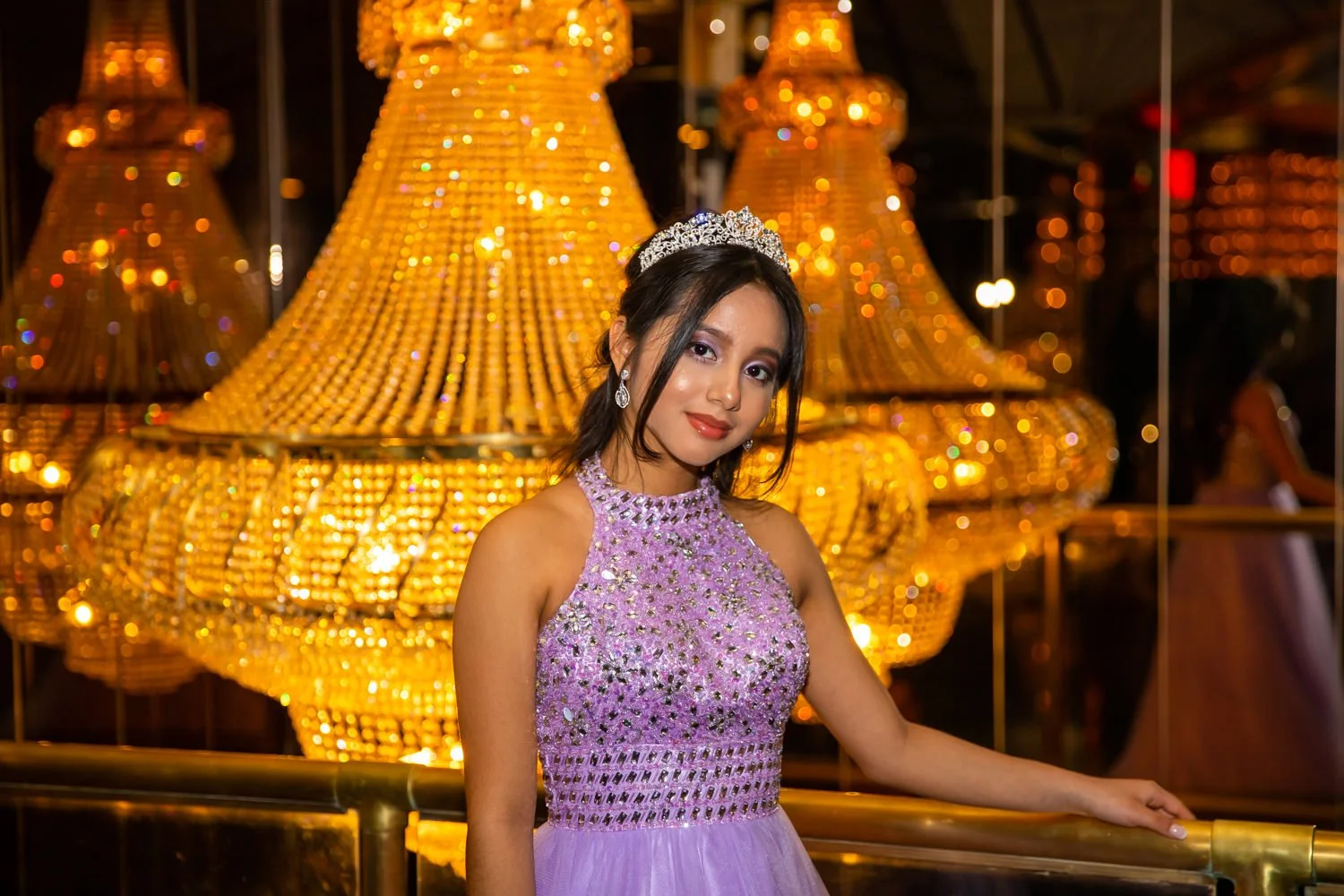 Young woman in lavender dress with jewelry and tiara, standing in front of large gold chandelier at a celebration or event.