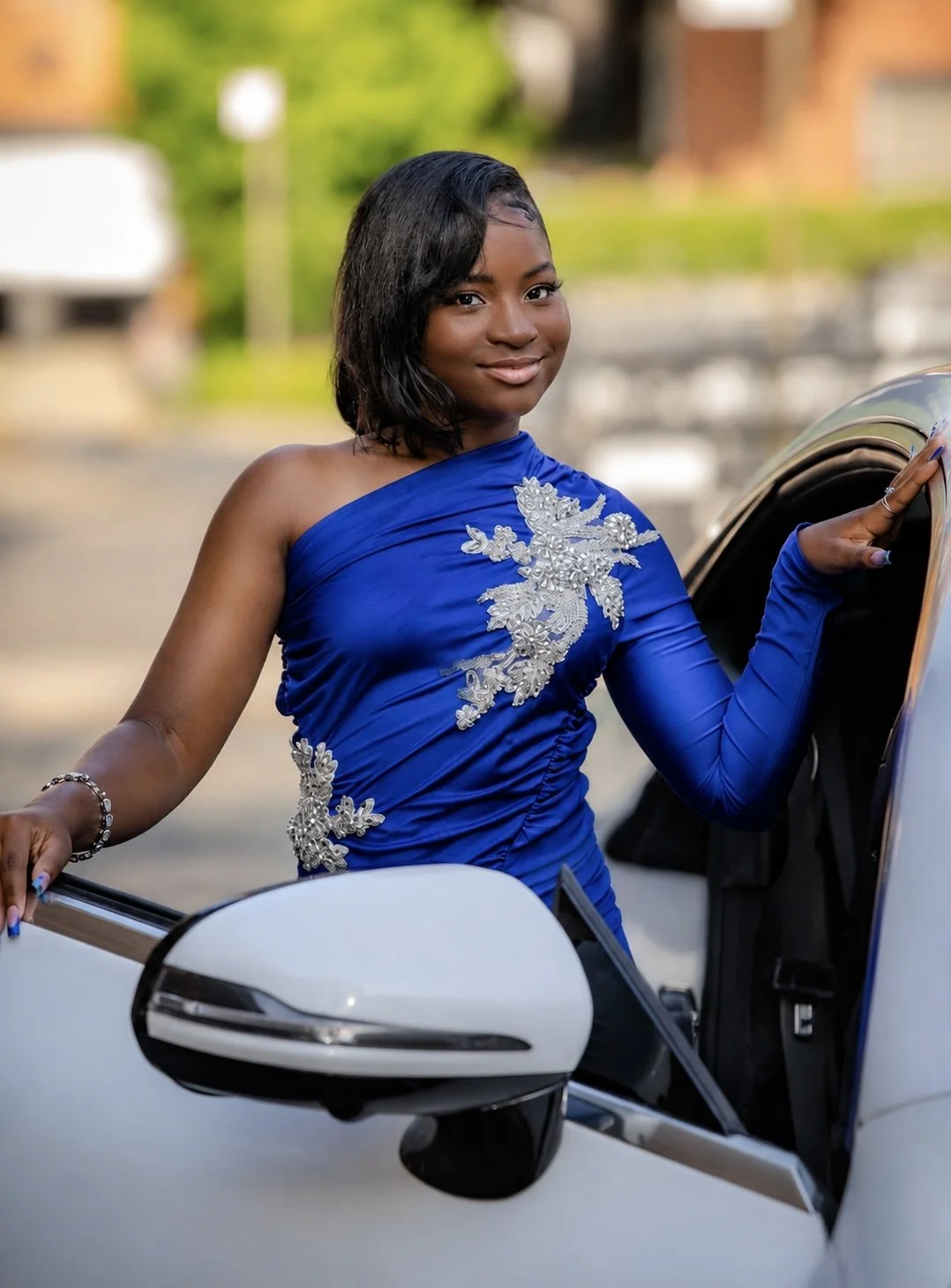 A young woman in a royal blue dress with silver embroidery, standing next to a white car, outdoors with blurred greenery in the background.