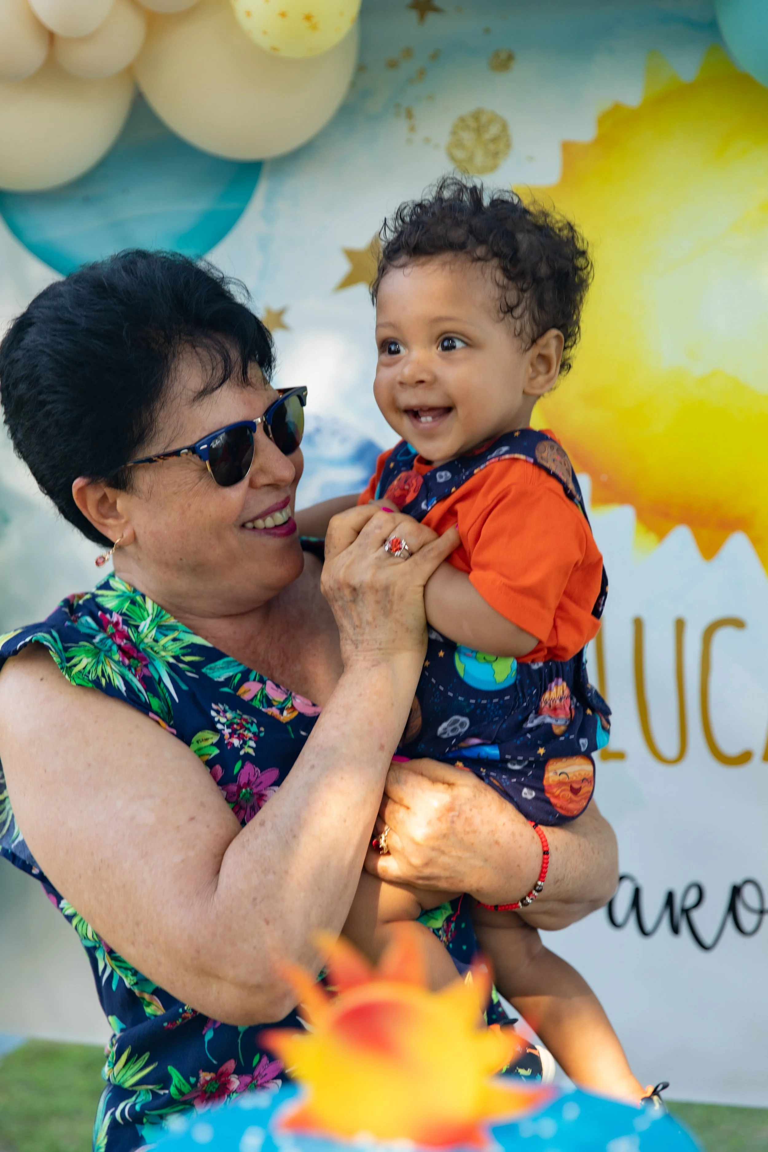 An elderly woman with short dark hair, wearing sunglasses and a colorful floral sleeveless dress, holding a smiling young boy with curly hair, dressed in an orange shirt with space-themed designs. They are outdoors with balloons and star decorations 