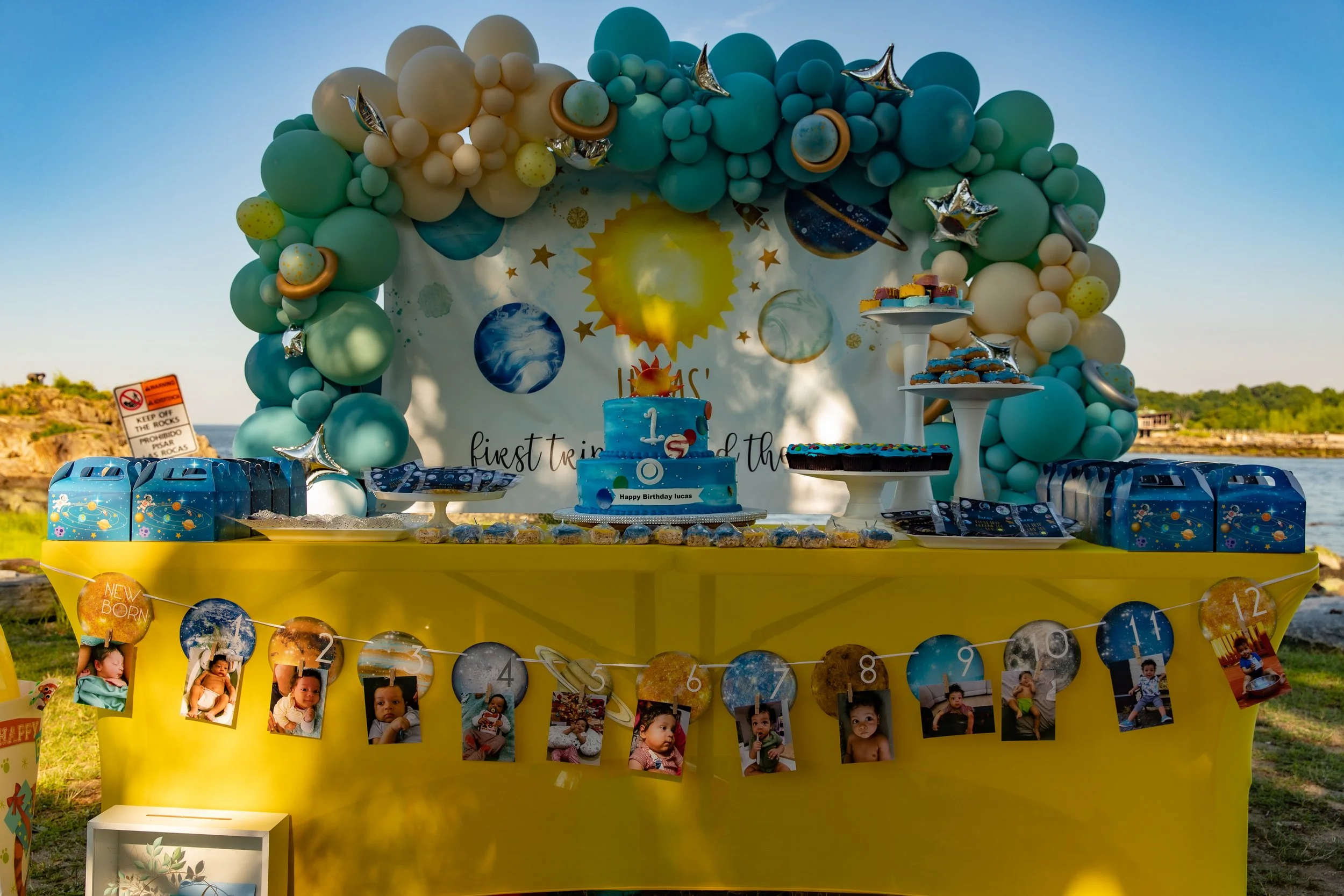 A colorful space-themed birthday party setup with a yellow table, decorated with photos of a child, a cake with space design, balloons in shades of green, blue, and beige, and space-themed decorations including planets and stars. An arch of balloons 
