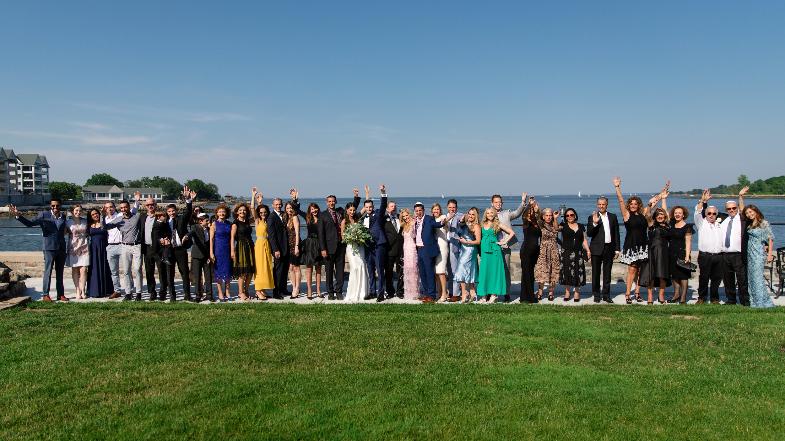 A large group of people posing outdoors near water, dressed in formal and colorful attire, with a cityscape and sailboats in the background.