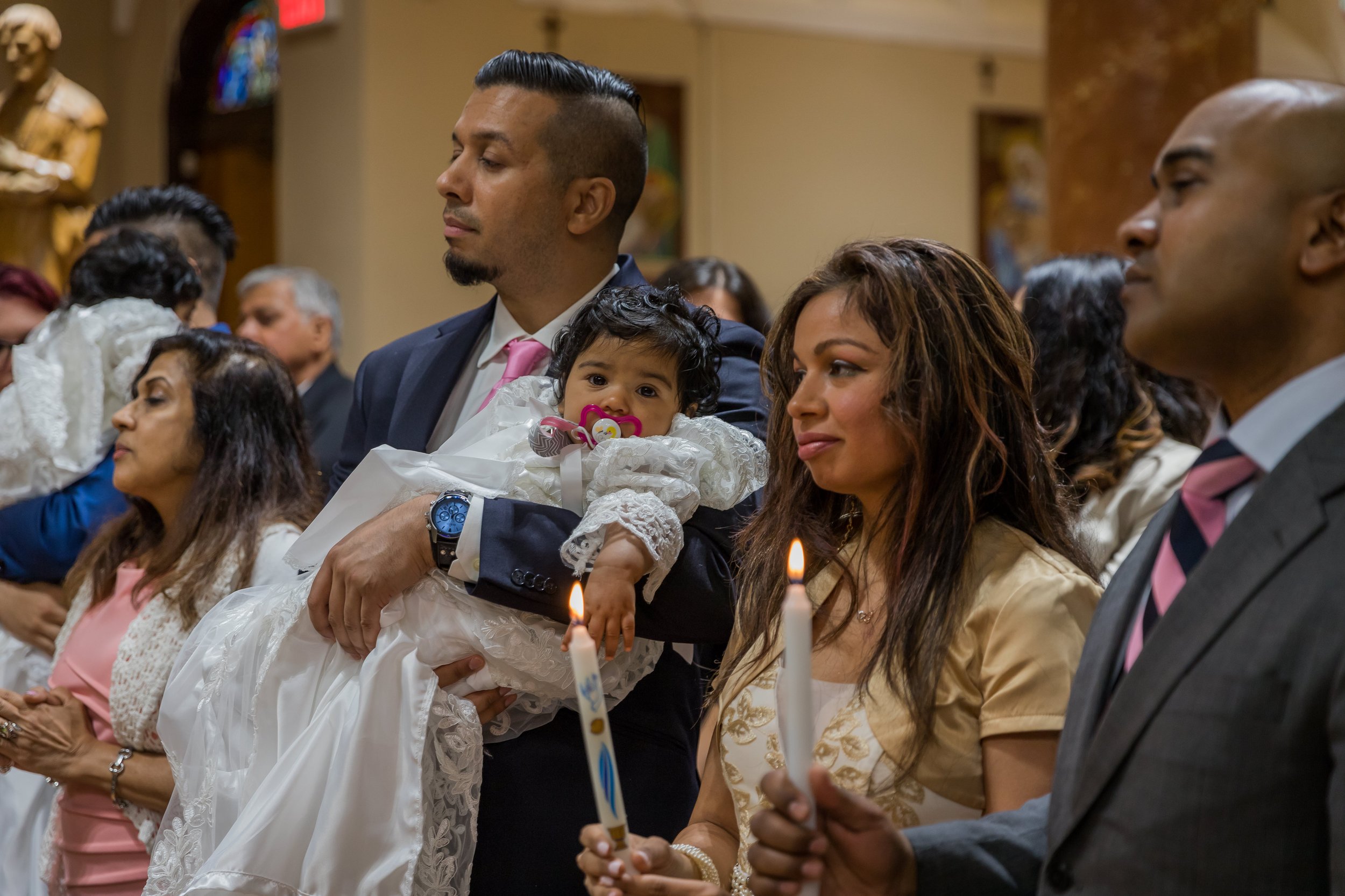 People attending a religious ceremony inside a church, holding candles and a young girl dressed in white with a pacifier, sitting on a man’s lap in front of a statue and stained glass window.