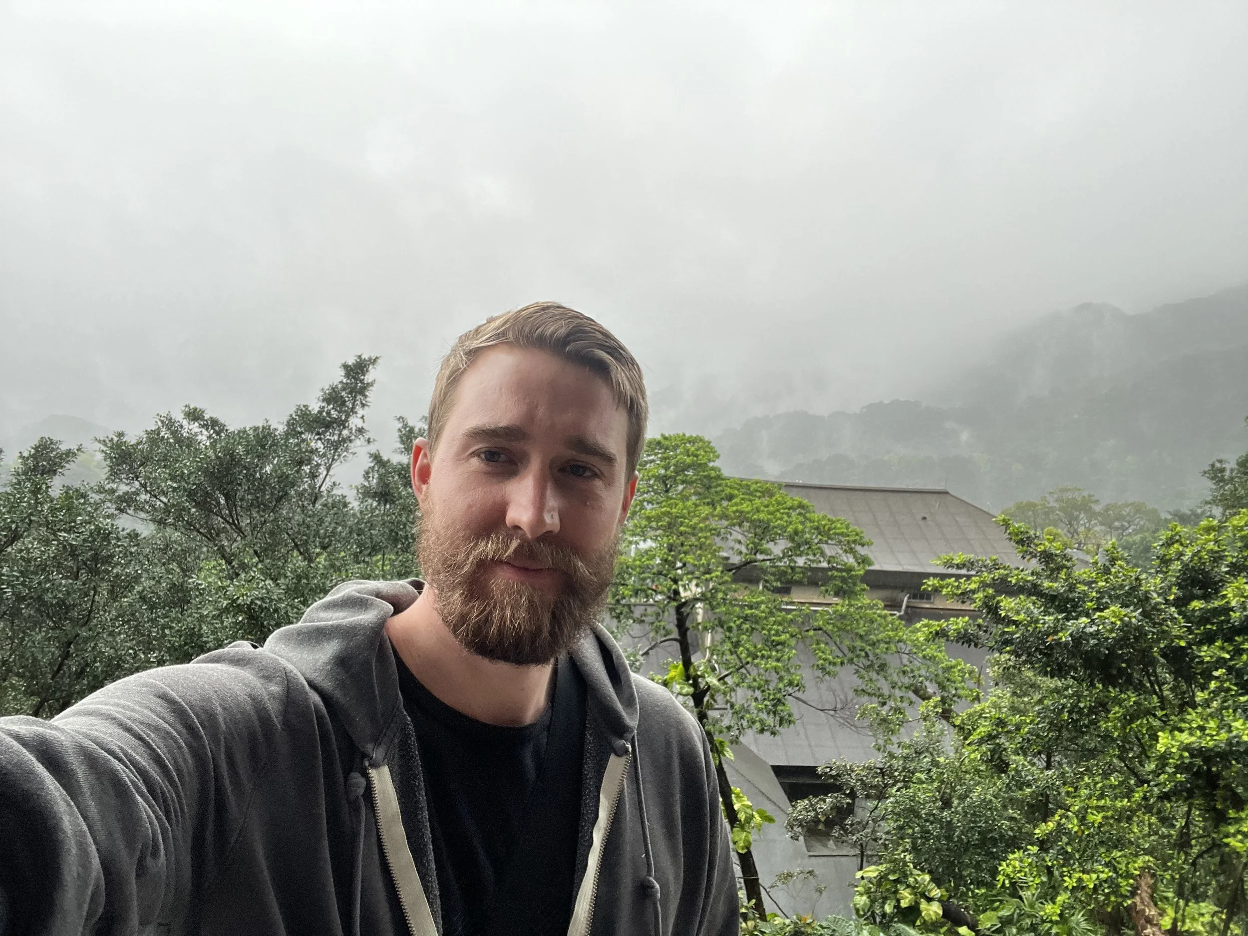 Man with short brown hair and a beard taking a selfie outdoors with trees and a house behind him on a cloudy day.