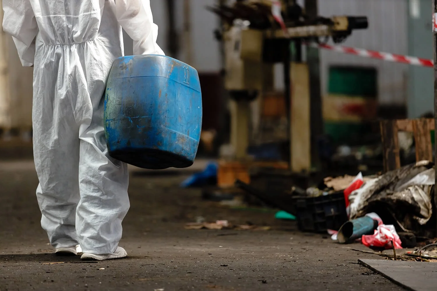 Person wearing a white protective suit holding a large blue bucket in an industrial or waste facility setting with scattered debris on the ground and cluttered workbenches in the background.