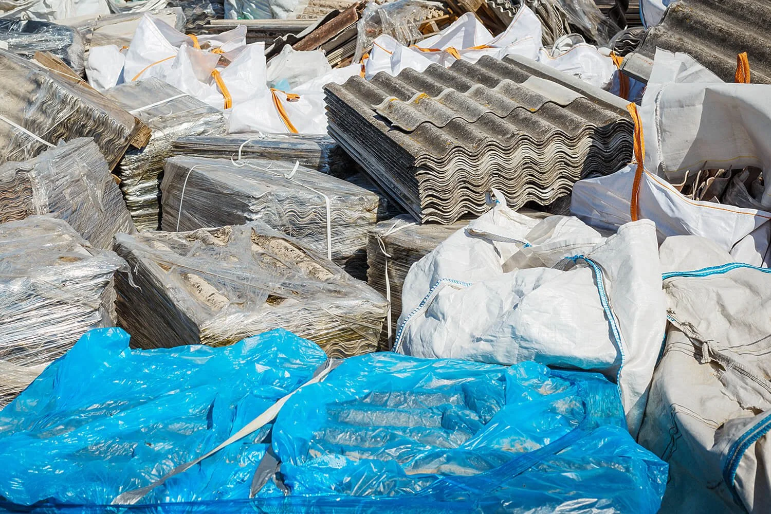 Piles of metallic roofing sheets, plastic bags, and bundled materials at a construction or scrap yard.