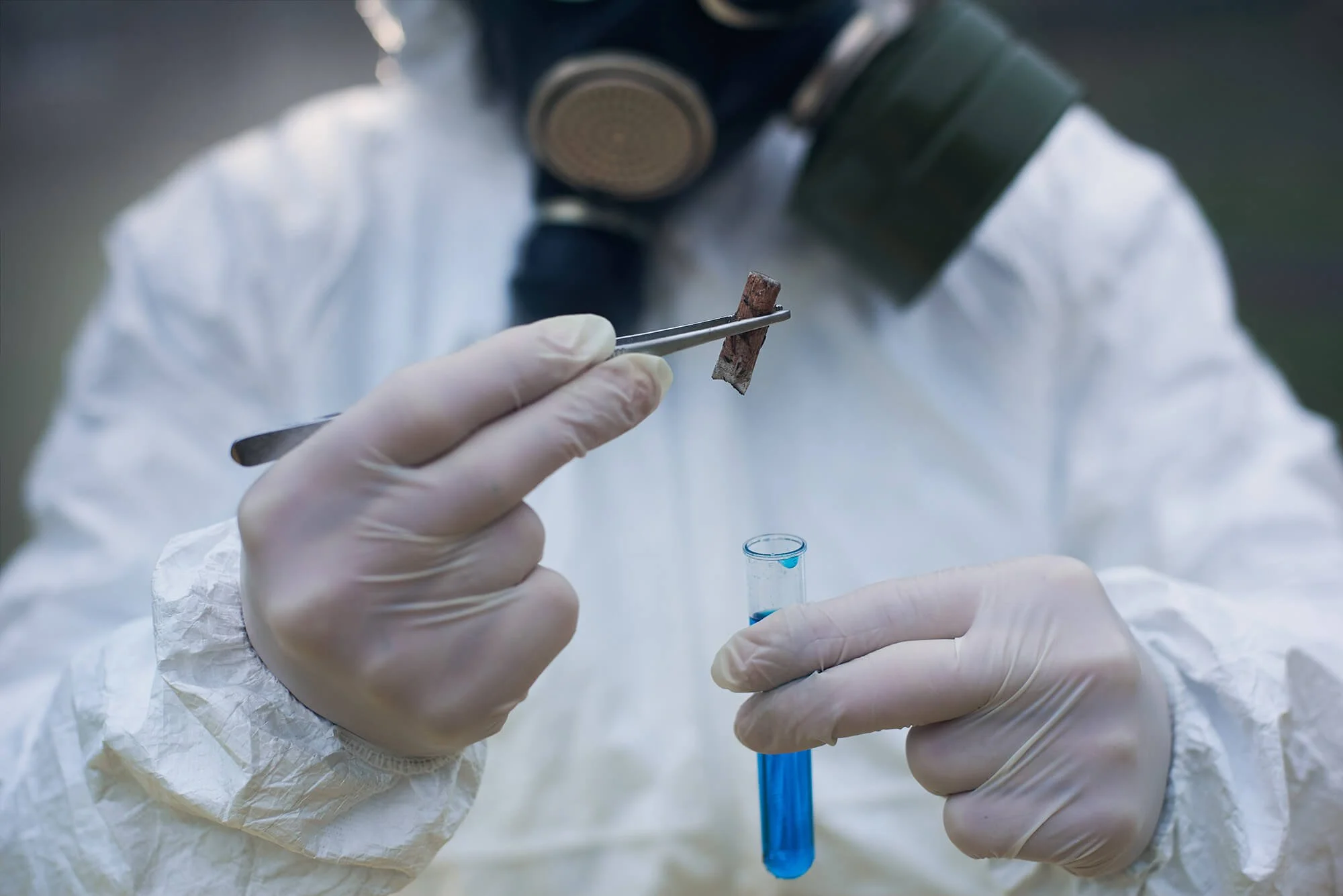 Scientist or researcher wearing gloves and a white protective suit using tweezers to handle a small sample of material above a test tube with blue liquid. A person wearing a mask and protective gear is visible in the background.