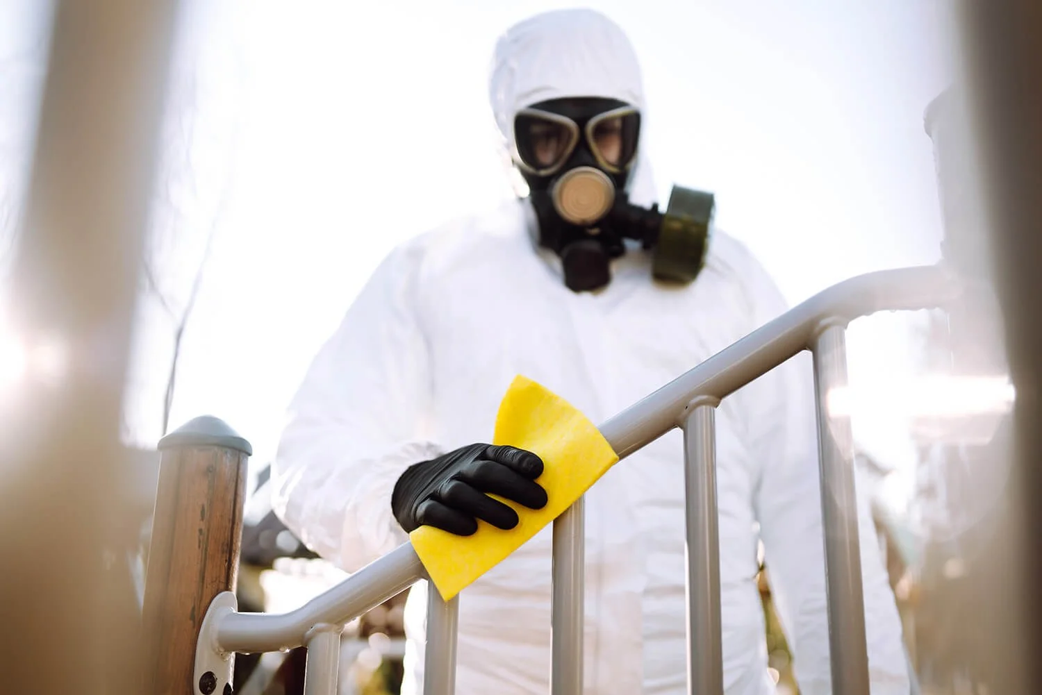 Person wearing a hazmat suit, gloves, and a gas mask cleaning a metal railing with a yellow sponge.