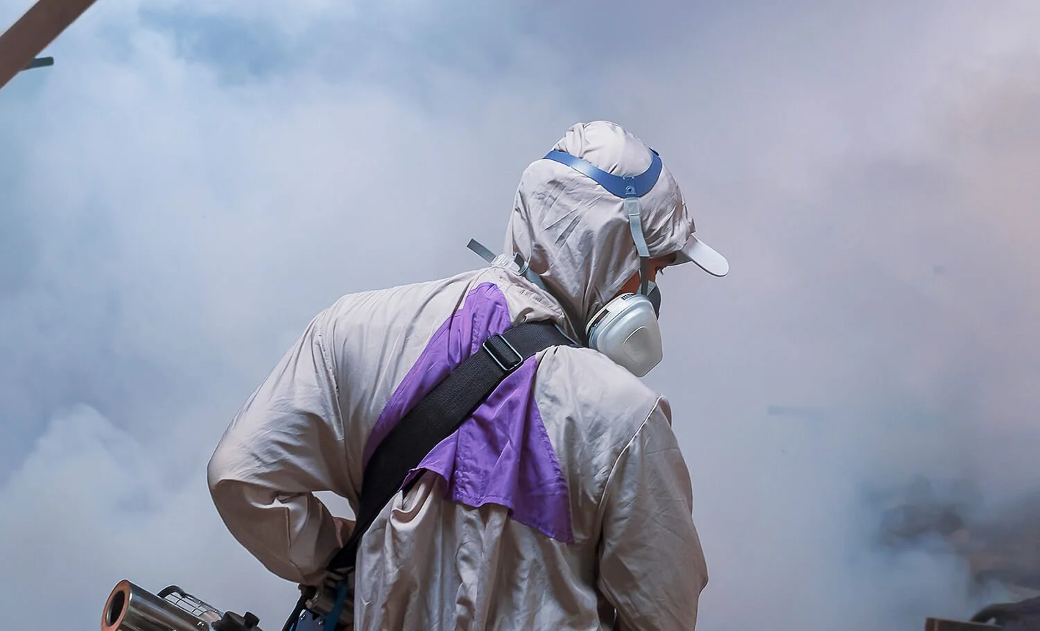 A person in protective gear sprays disinfectant amidst thick smoke or fog.