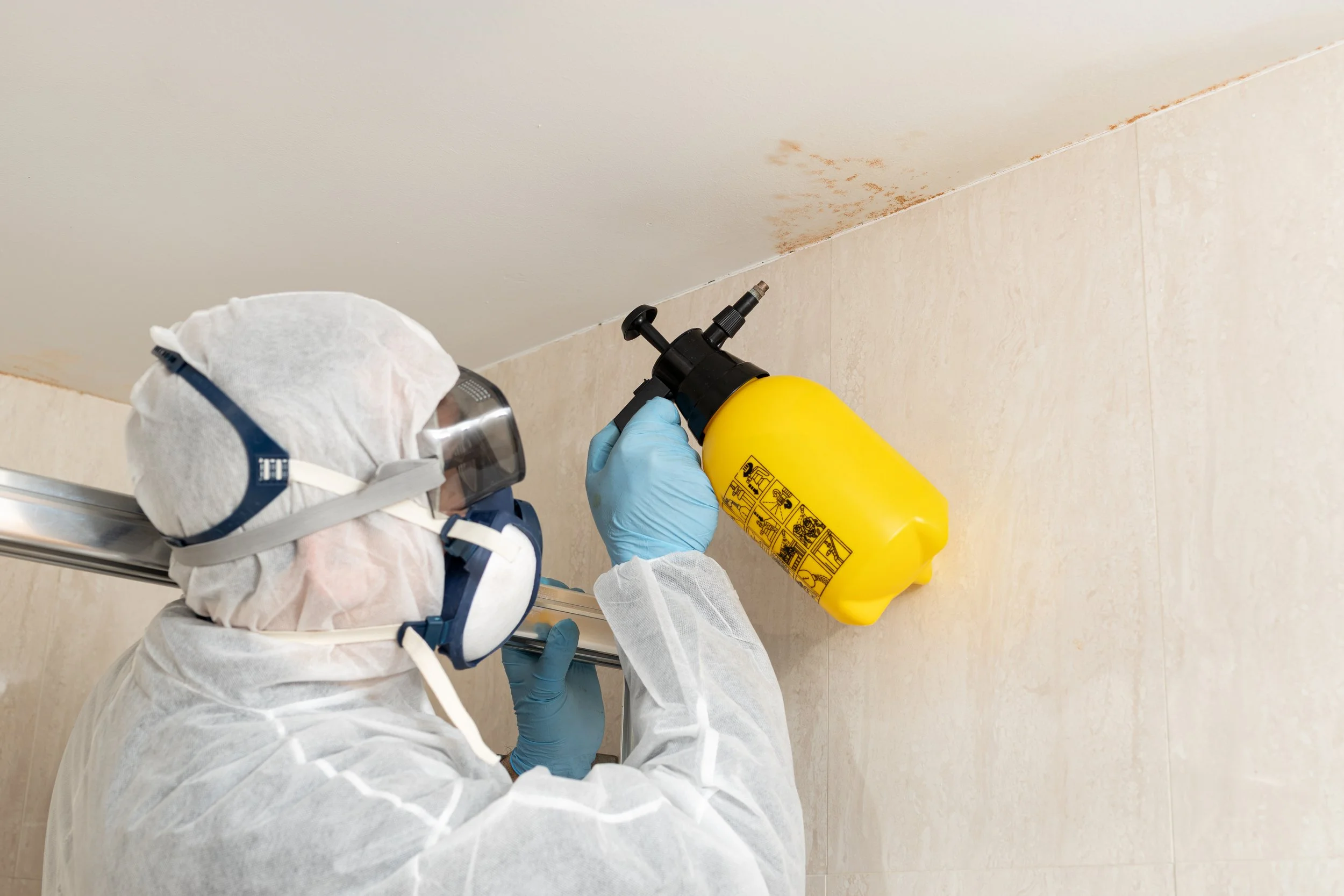 Person wearing protective gear using a yellow spray bottle to apply a cleaning or disinfectant solution to a ceiling.