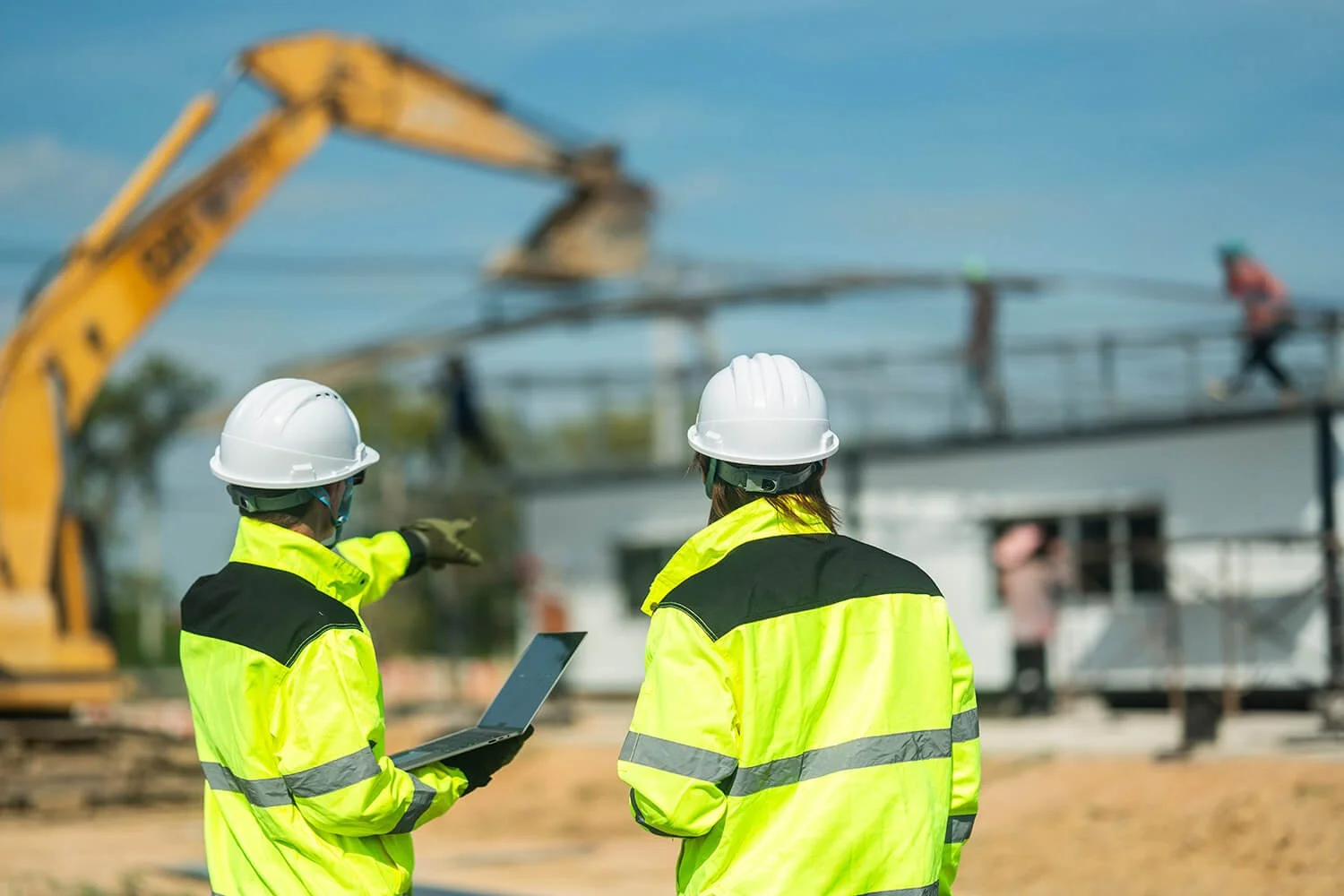 Two construction workers in yellow safety jackets and white helmets on a construction site, with one holding a laptop and one pointing toward the structure.