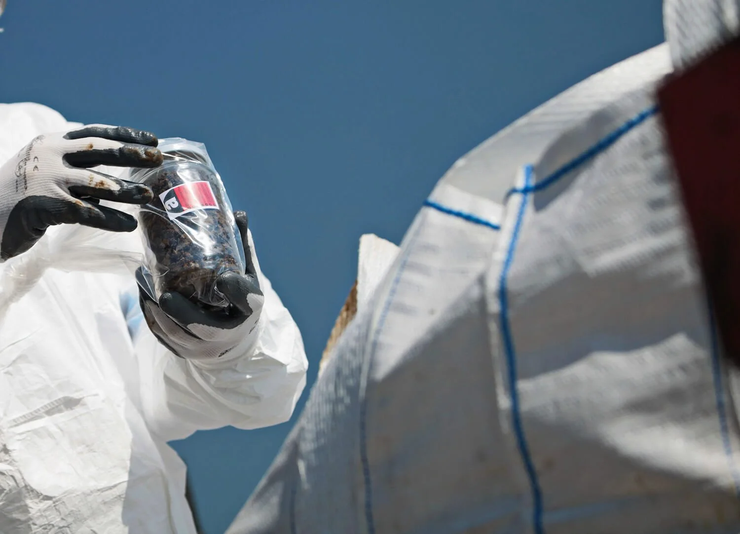 A person in white protective suit and gloves holds a small plastic bag with dark objects inside, possibly research samples, with a clear blue sky in the background.