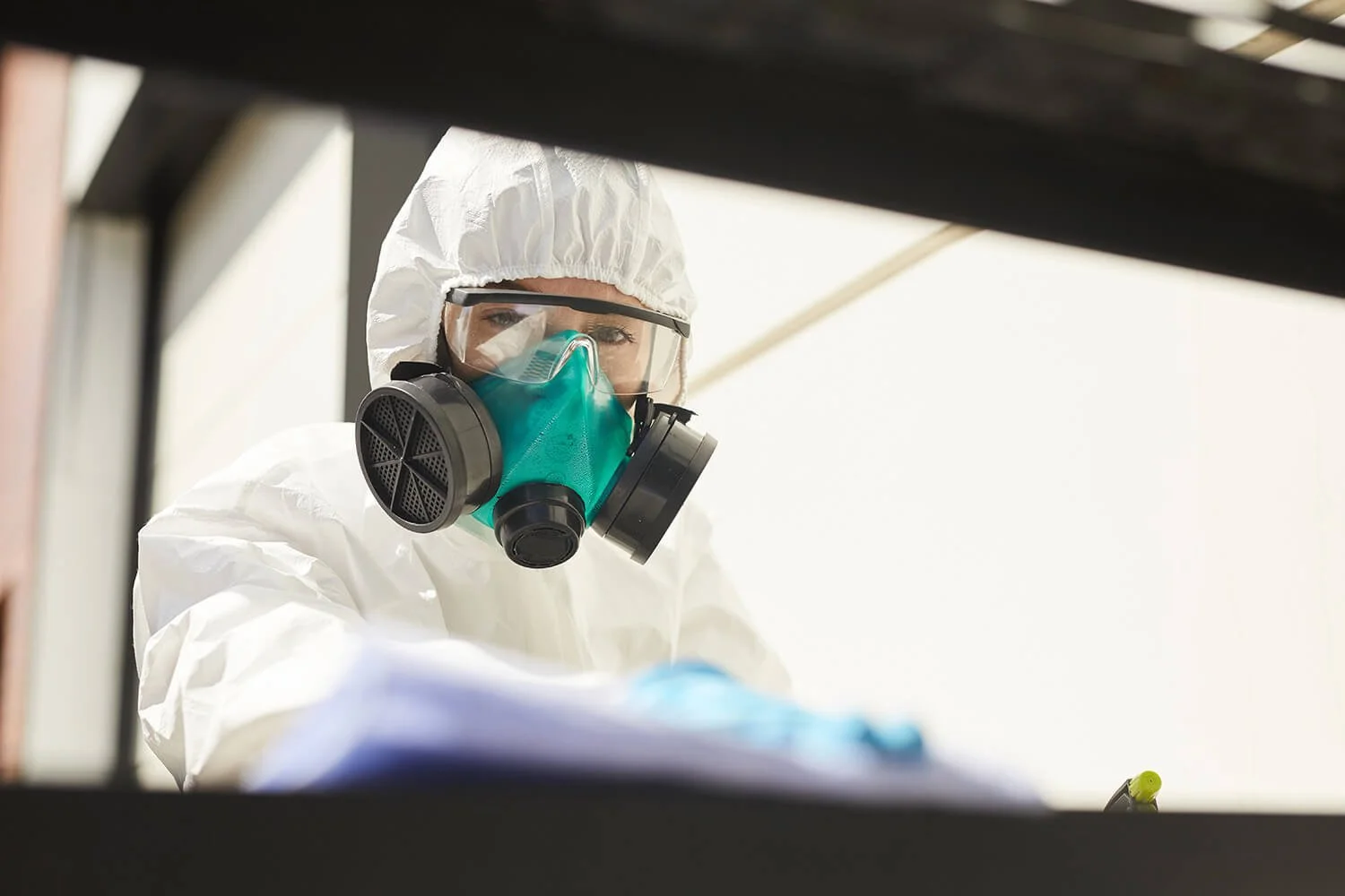 A person dressed in full protective gear, including a white hazmat suit, goggles, and a green respirator mask, working in a laboratory or industrial setting.