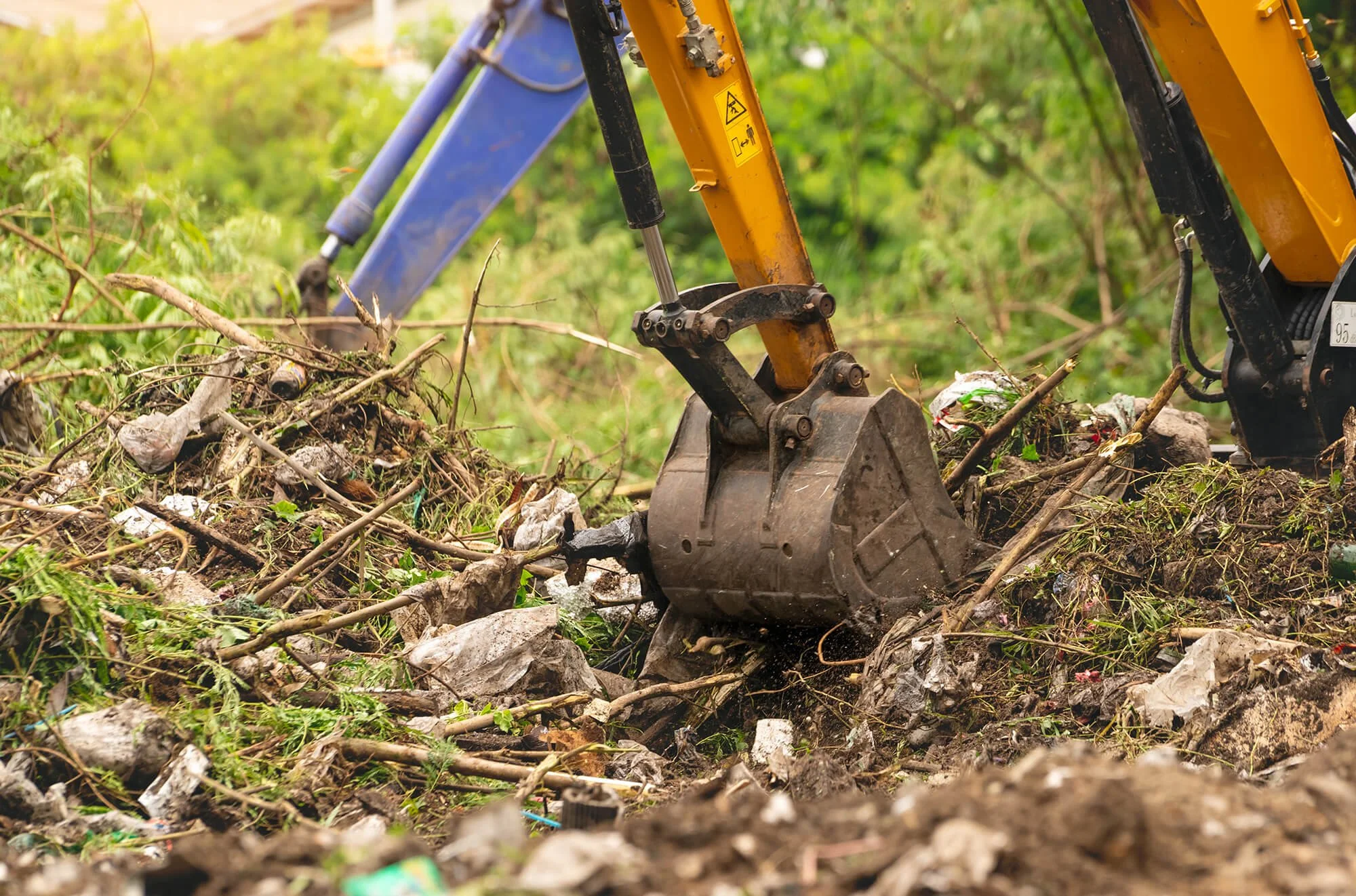 Part of a small excavator digging through trash and debris in a green outdoor environment.