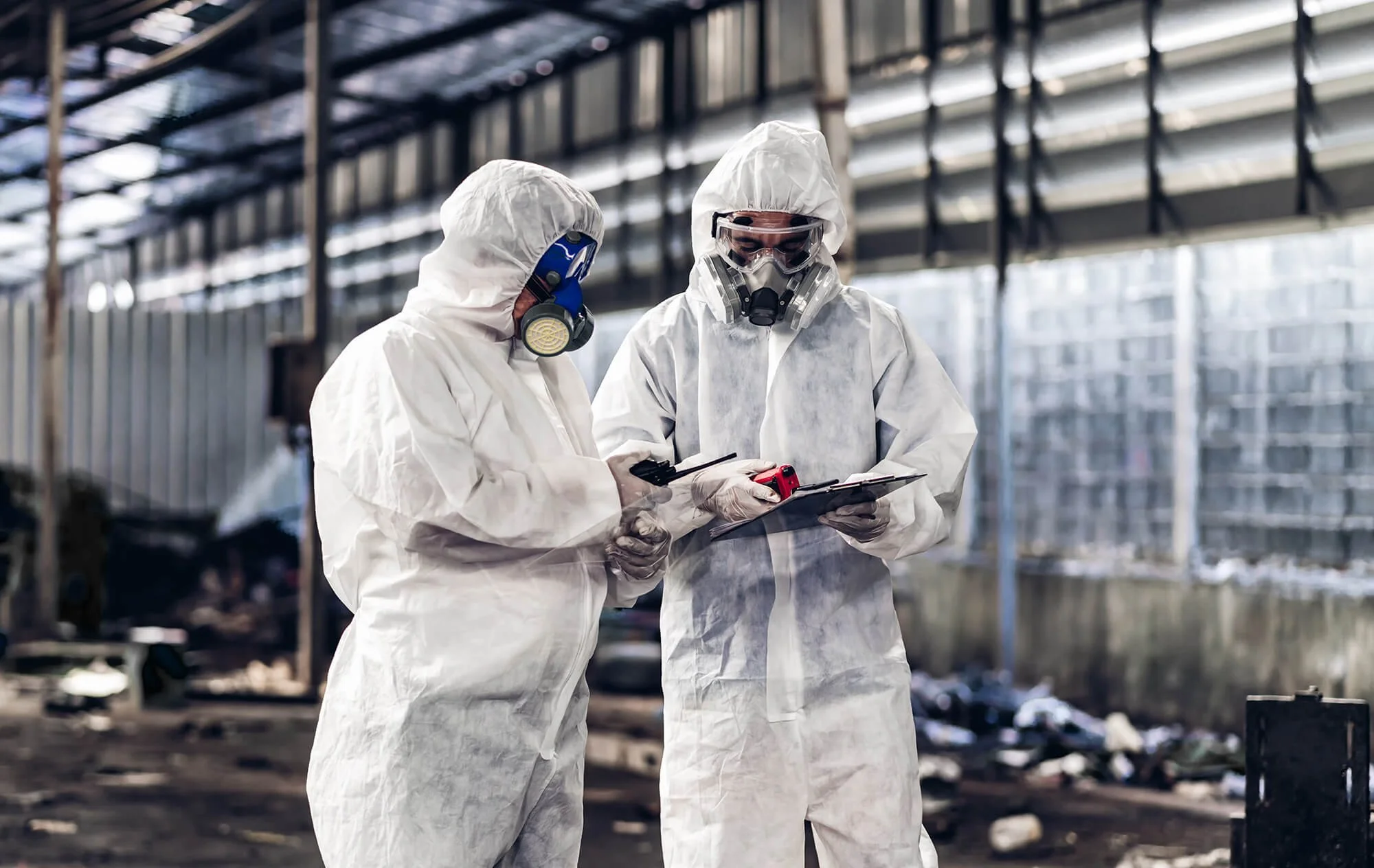 Two workers in full protective suits, masks, and goggles standing in an industrial or construction setting, reviewing information on a clipboard and a mobile device.