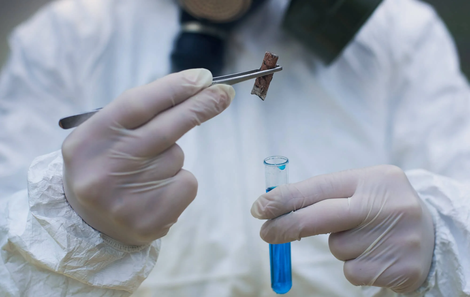 A scientist wearing white gloves is holding a test tube with blue liquid and using forceps to place a small wooden piece into a test tube.