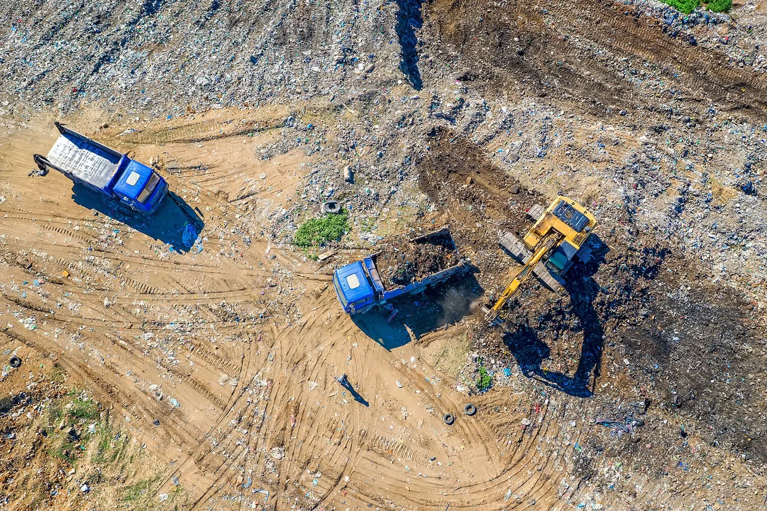 Aerial view of a landfill site with two trucks and a bulldozer moving debris and garbage on a dirt area.