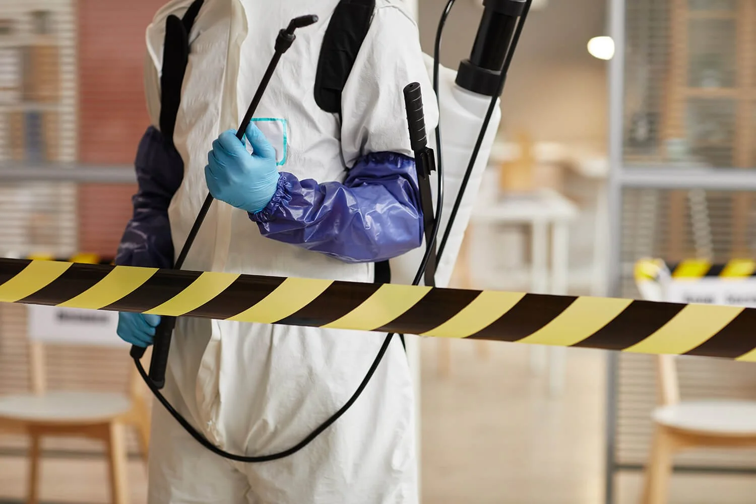 A person in protective gear holding a sprayer, standing behind yellow and black caution tape in a warehouse or storage area.