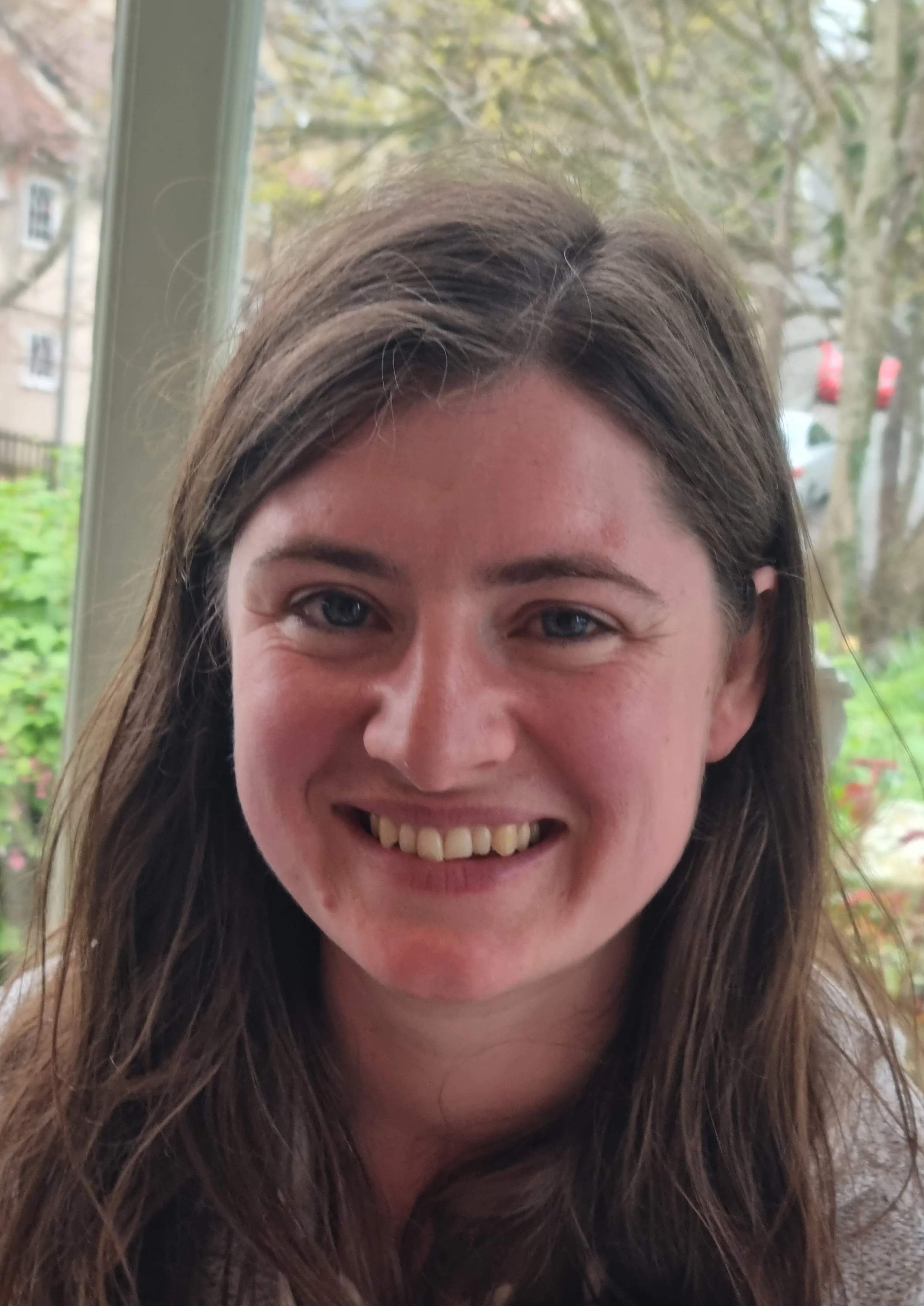Close-up of a young woman's face with a big smile, showing her teeth, with long brown hair and blue eyes, outdoors with trees and cars in the background.