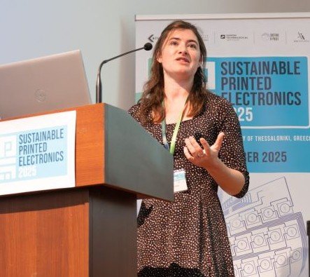 Woman speaking at a conference podium with a laptop, wearing a badge, and gesturing with her hand, with a blue and white conference banner in the background.