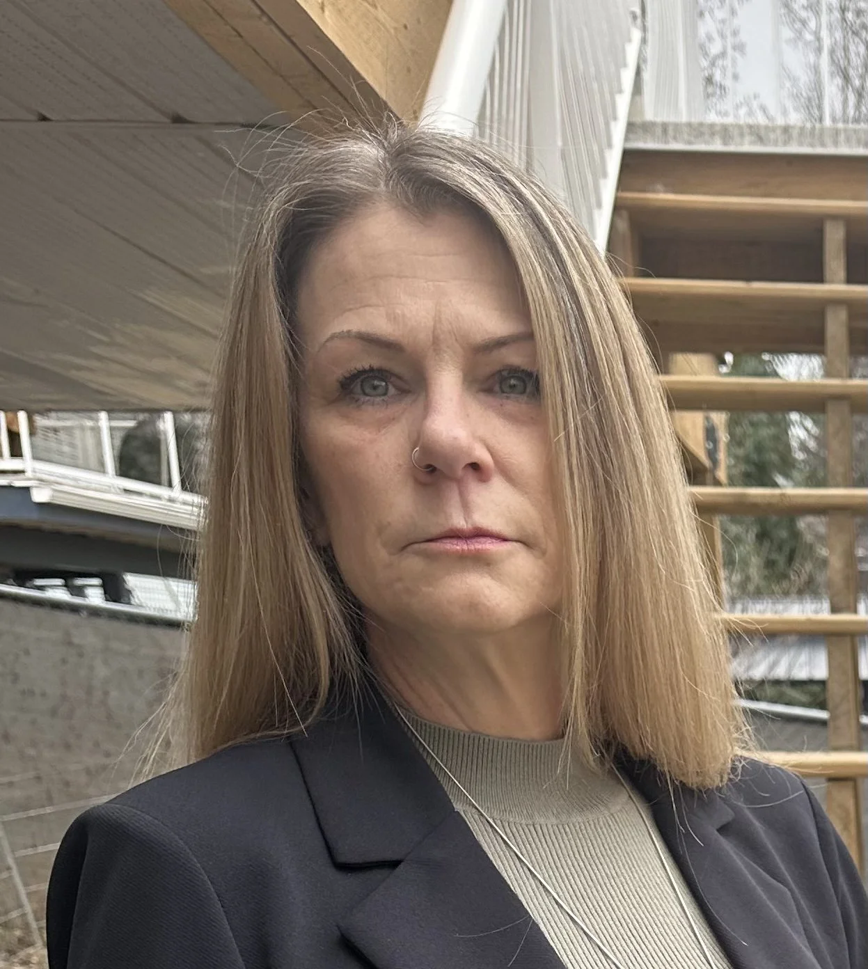 A woman with blonde hair and a serious expression, wearing a black blazer and a necklace, standing outdoors near a wooden staircase and a cabin or house.