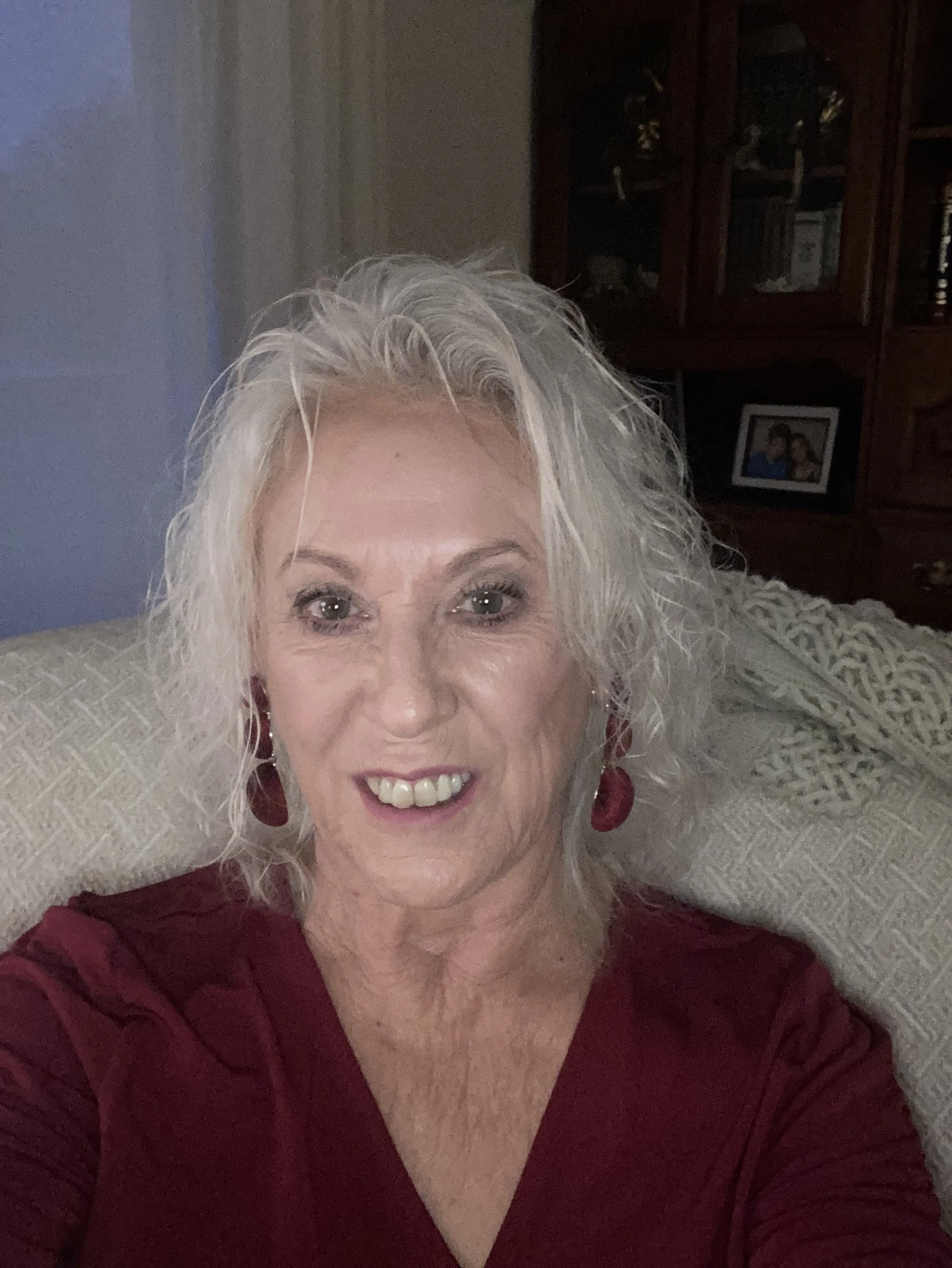 Close-up of a smiling older woman with curly gray hair, wearing a burgundy top and matching earrings, lying on a light-colored textured surface with a wooden cabinet and family photos in the background.