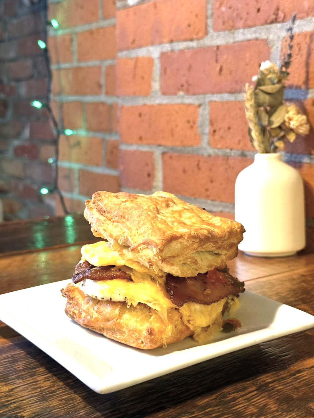 A plate with a breakfast sandwich of fried eggs, bacon, and a croissant on a wooden table, with a brick wall and a white vase with dried flowers in the background.