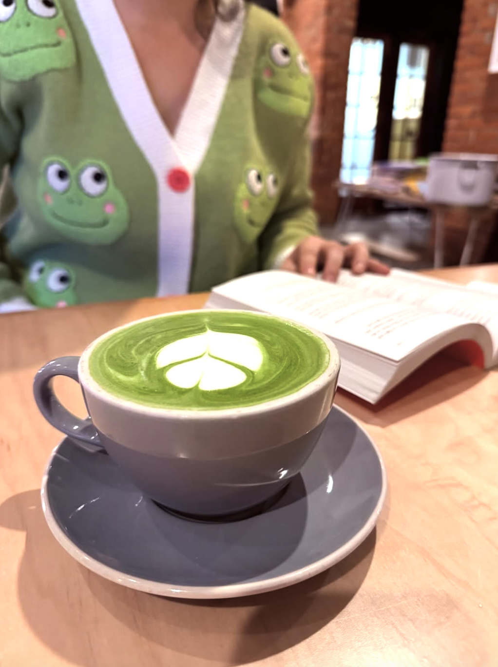 A cup of green matcha latte with latte art in the shape of a four-leaf clover on a saucer, placed on a wooden table. In the background, a person in a green sweater with frog patterns is reading a book in a cozy cafe with brick walls and windows.