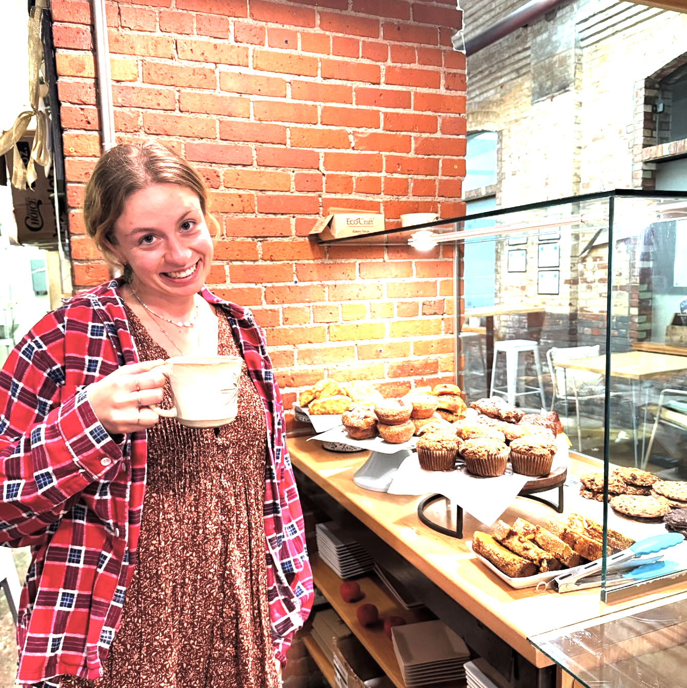 A young woman with a smile, wearing a red plaid shirt, holding a mug, standing next to a display of baked goods like muffins, cookies, and bars inside a cafe with a brick wall and glass display case.
