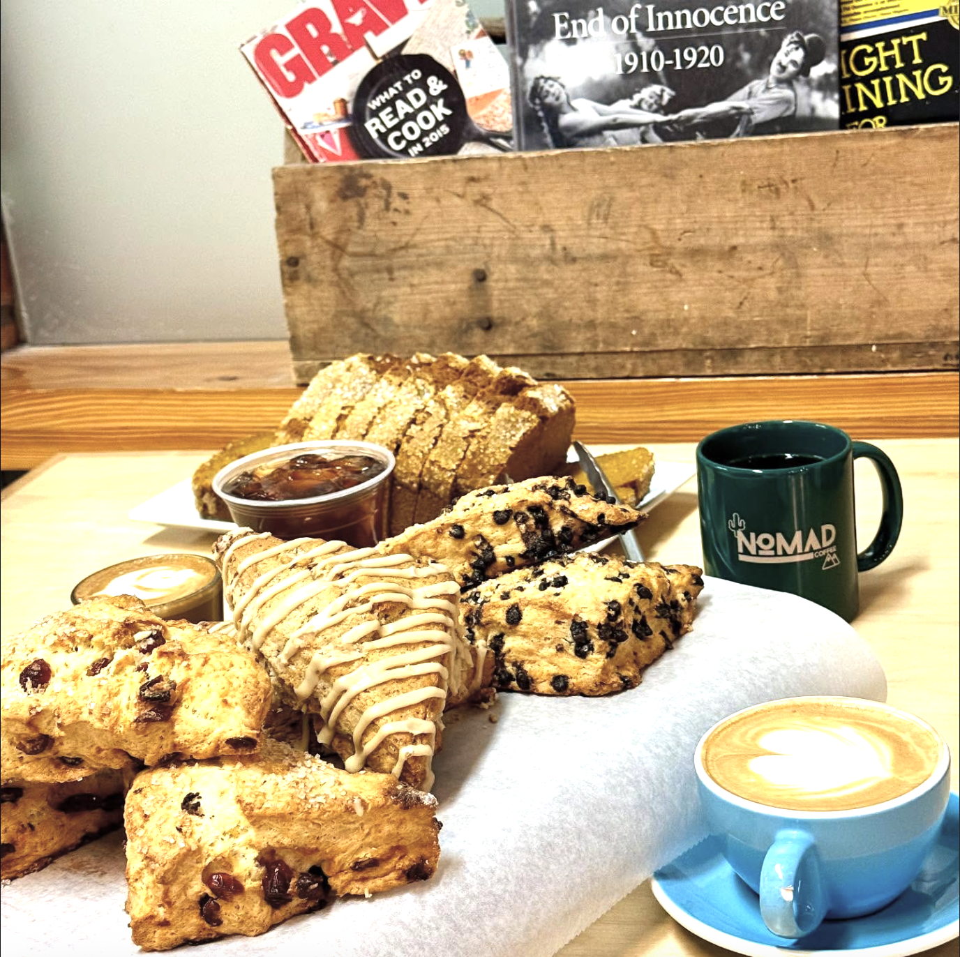 Plate of assorted baked goods including cookies, biscotti, and scones with chocolate chips, along with cups of coffee and a small container of jam on a table.