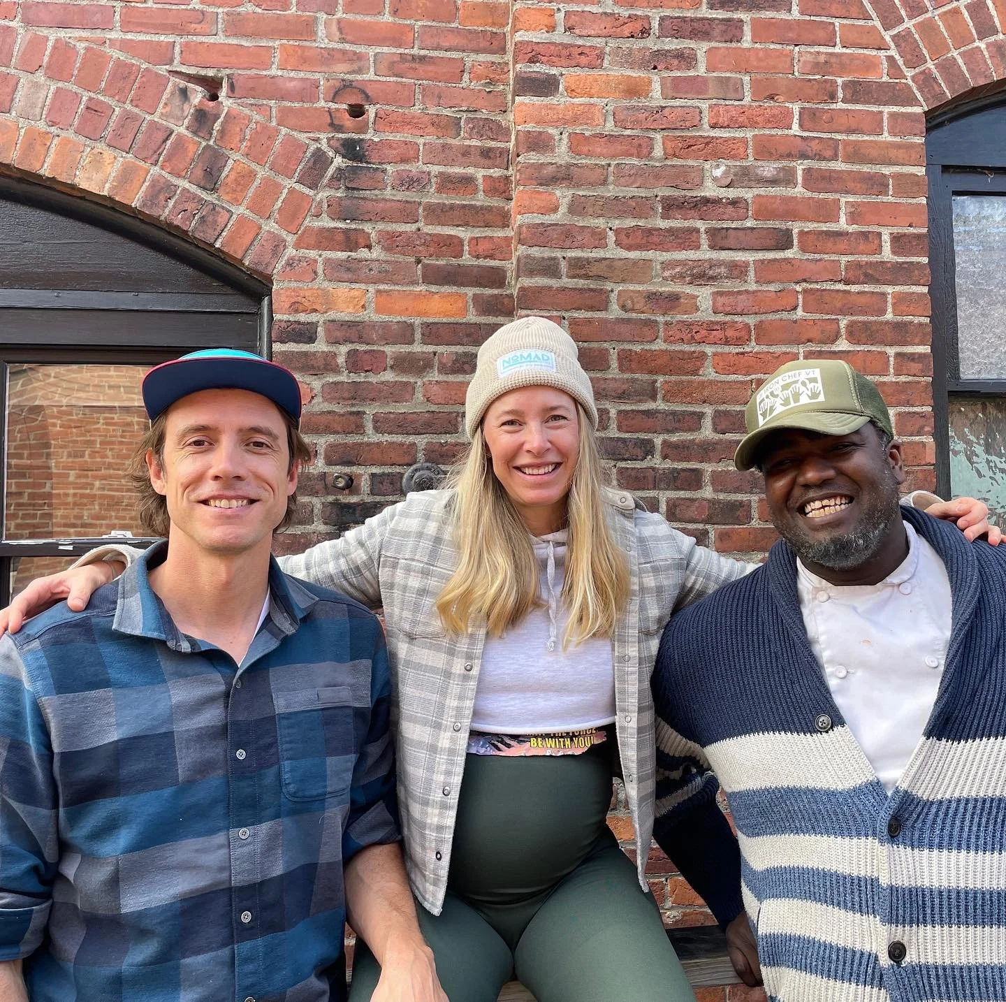 Three people smiling and sitting together outdoors in front of a brick wall, with two windows behind them.