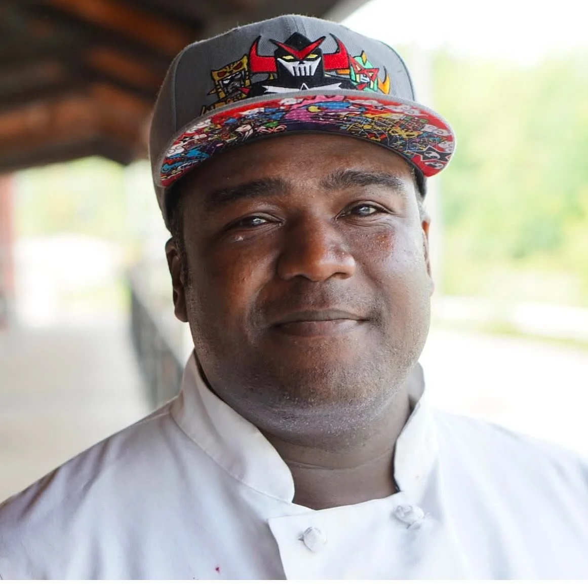 A man wearing a colorful baseball cap with cartoon characters and a white chef's coat, standing outdoors with a blurred background.