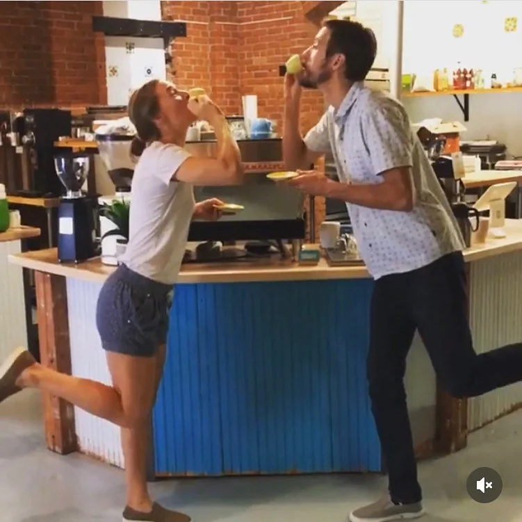 A woman and man are alternately pretending to eat ice cream cones in a cafe. The woman is on the left, standing on one leg, while the man is on the right, holding a plate and leaning toward her.