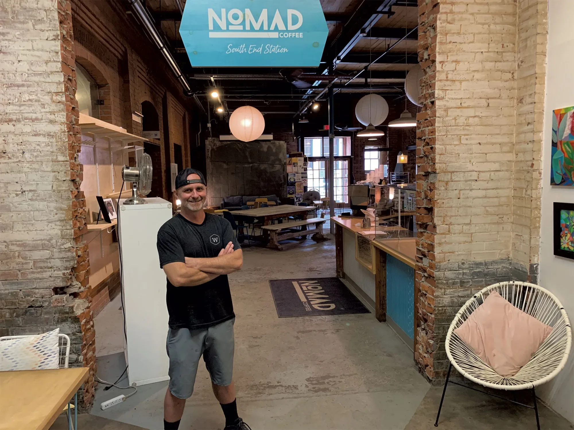 A man smiling and standing with crossed arms inside a coffee shop called Nomad Coffee, South End Station, with exposed brick walls and hanging paper lantern lights.