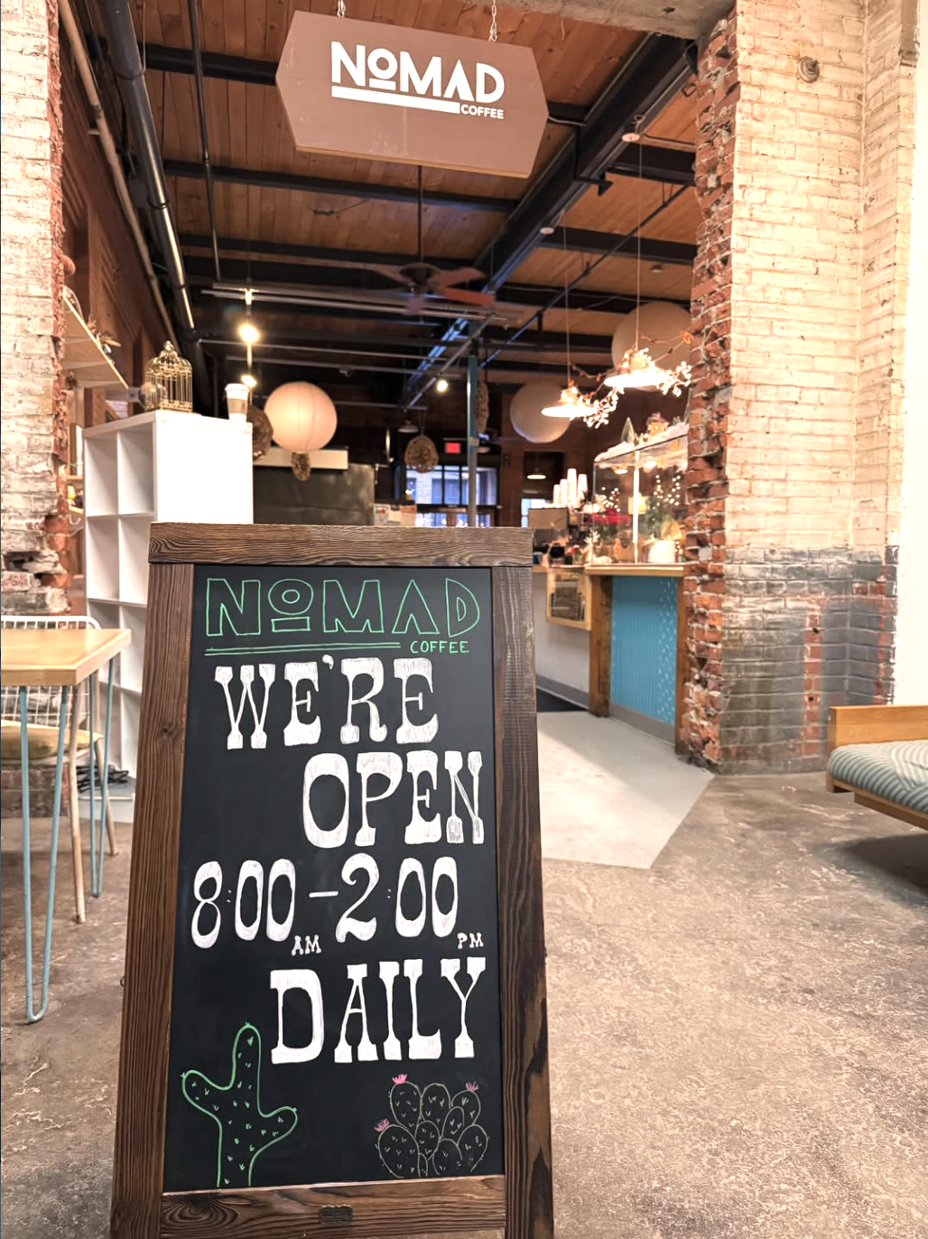 The image shows the interior of a coffee shop named Nomad Coffee. There is a blackboard sign in the foreground displaying the shop's opening hours, 8:00 AM to 2:00 PM daily, with decorative drawings of a cactus and a cluster of prickly pears. The shop has exposed brick walls, wooden accents, and warm lighting, creating a cozy atmosphere.