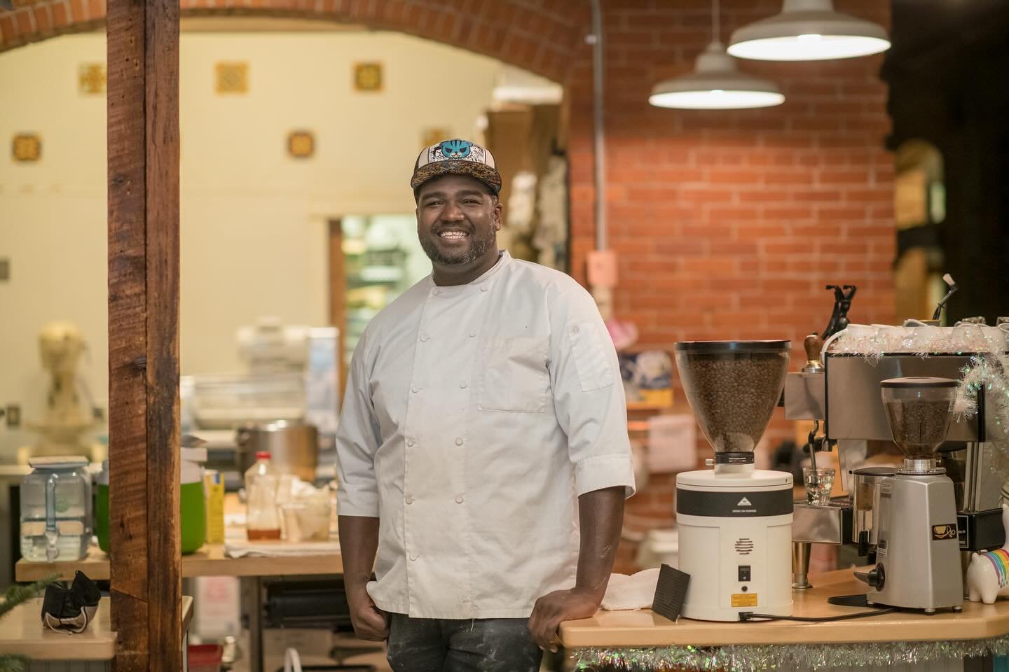 A smiling chef in a white uniform and patterned cap standing in a cozy kitchen with brick and wood accents, surrounded by coffee grinders and kitchen equipment.