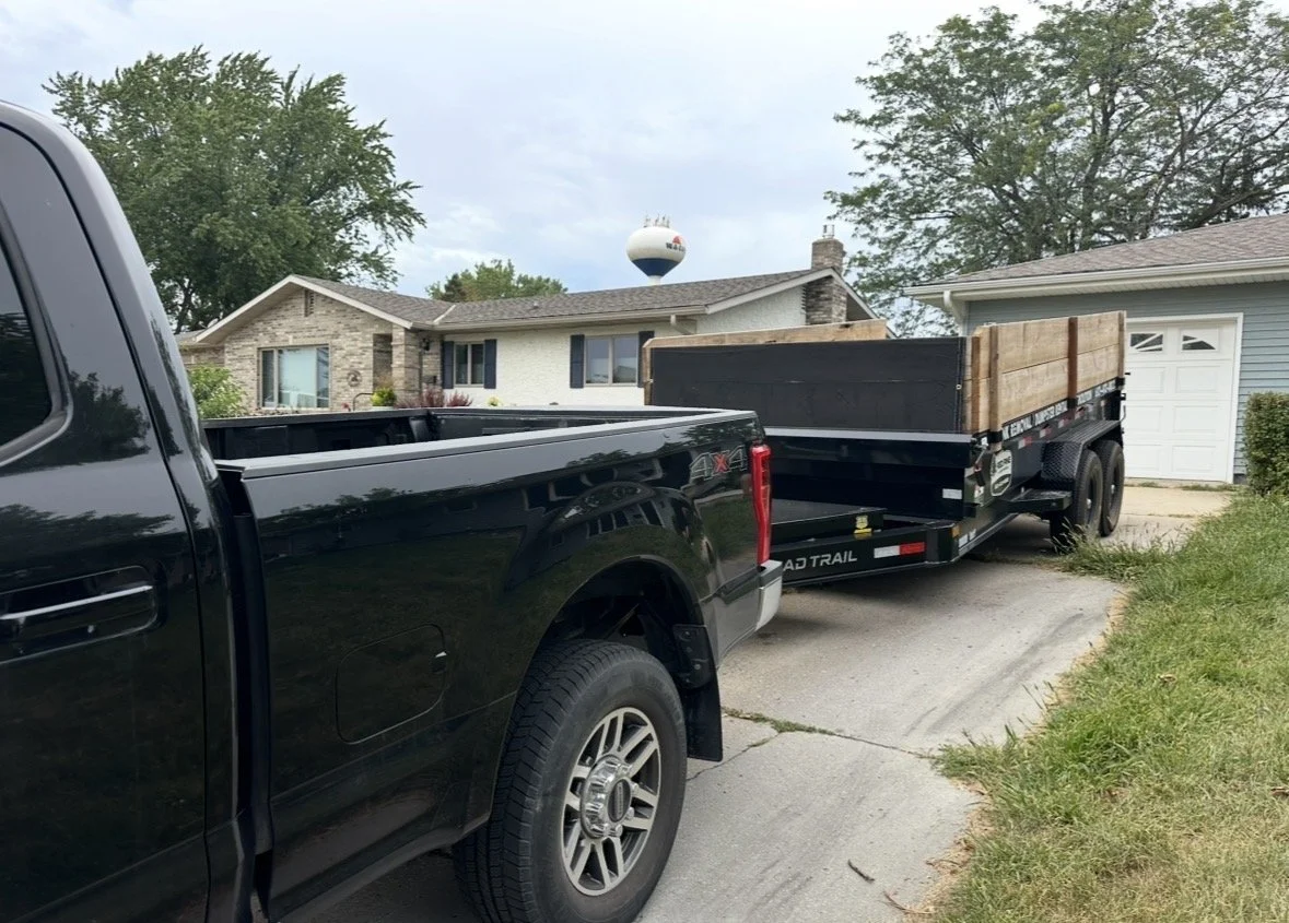 A black pickup truck towing a dump trailer with a wooden side panel parked in a residential driveway.