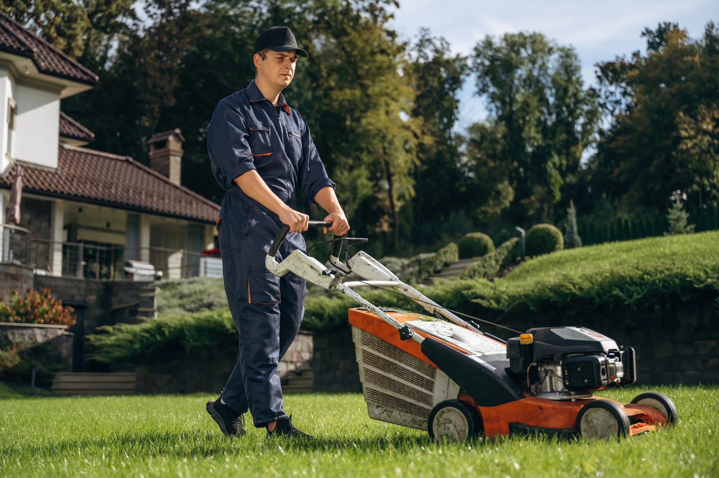 A man wearing a navy worker's jumpsuit and a black cap is mowing a lush green lawn with a large, orange and white walk-behind lawn mower.