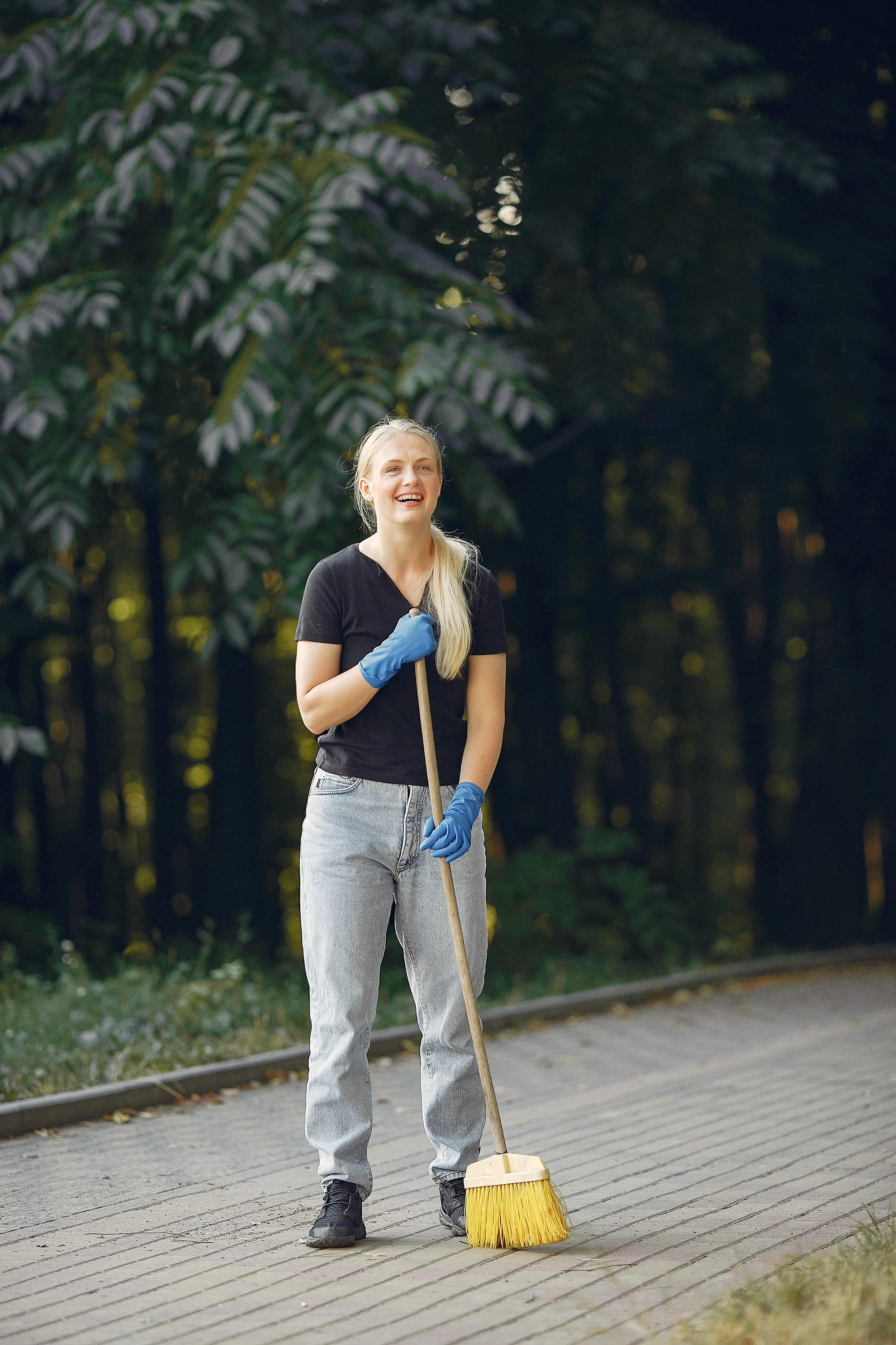 A young woman with blonde hair, wearing black t-shirt, grey pants, and blue gloves, is smiling while holding a broom on a paved walkway in a park or garden with lush green trees in the background.