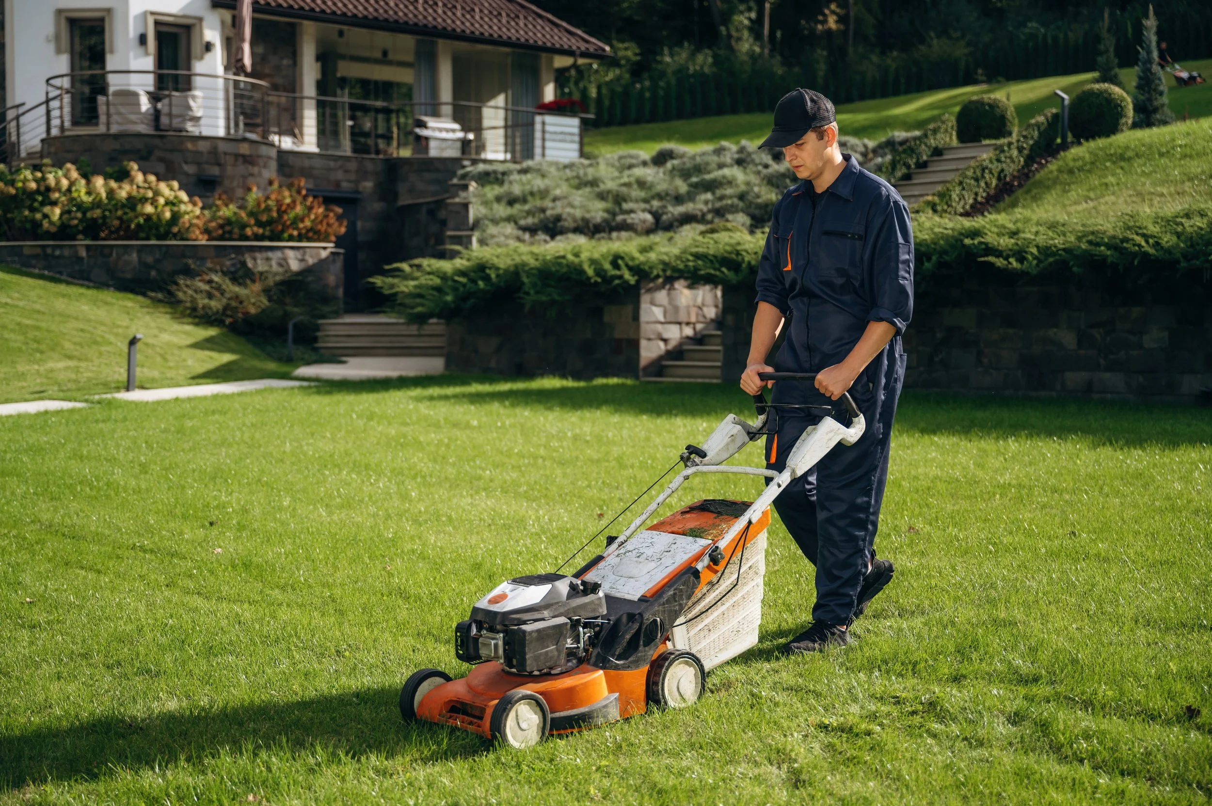 A man wearing a dark uniform and cap is cutting grass with a riding lawn mower on a well-maintained lawn in front of a house with multiple steps and plants.