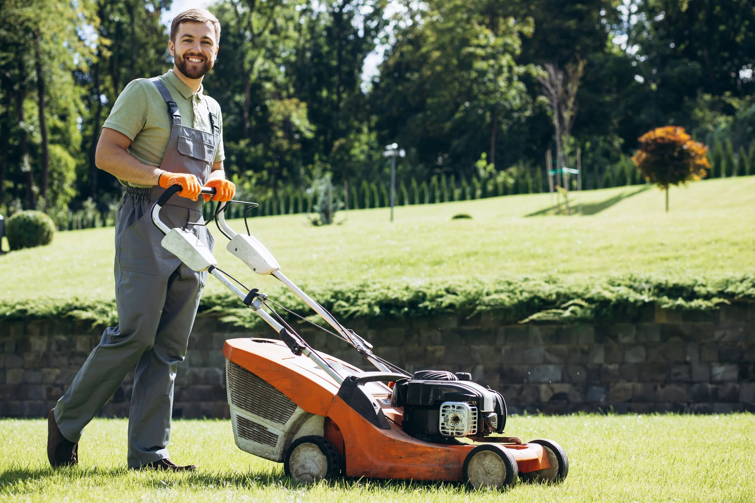 A man with a beard and brown hair, smiling, wearing a green polo shirt, grey overalls, and orange gloves, standing on green grass with a lawnmower in a park-like setting with trees and bushes.