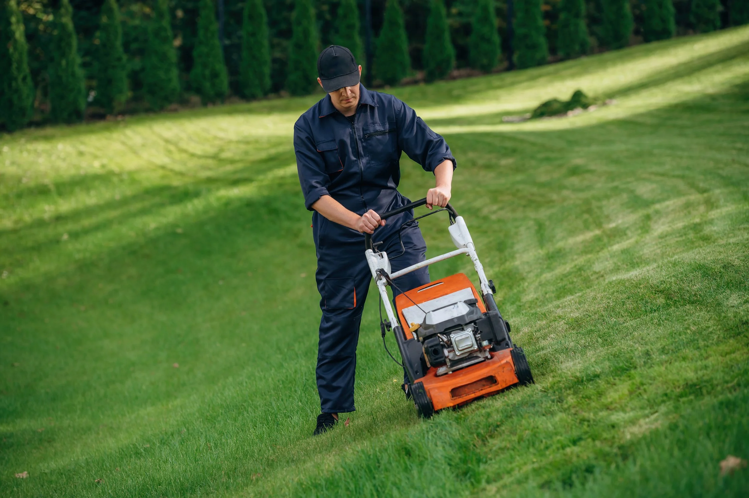 A man wearing a navy blue jumpsuit and black cap is mowing the lawn with an orange and black push lawn mower on a green grassy yard, with trees in the background.