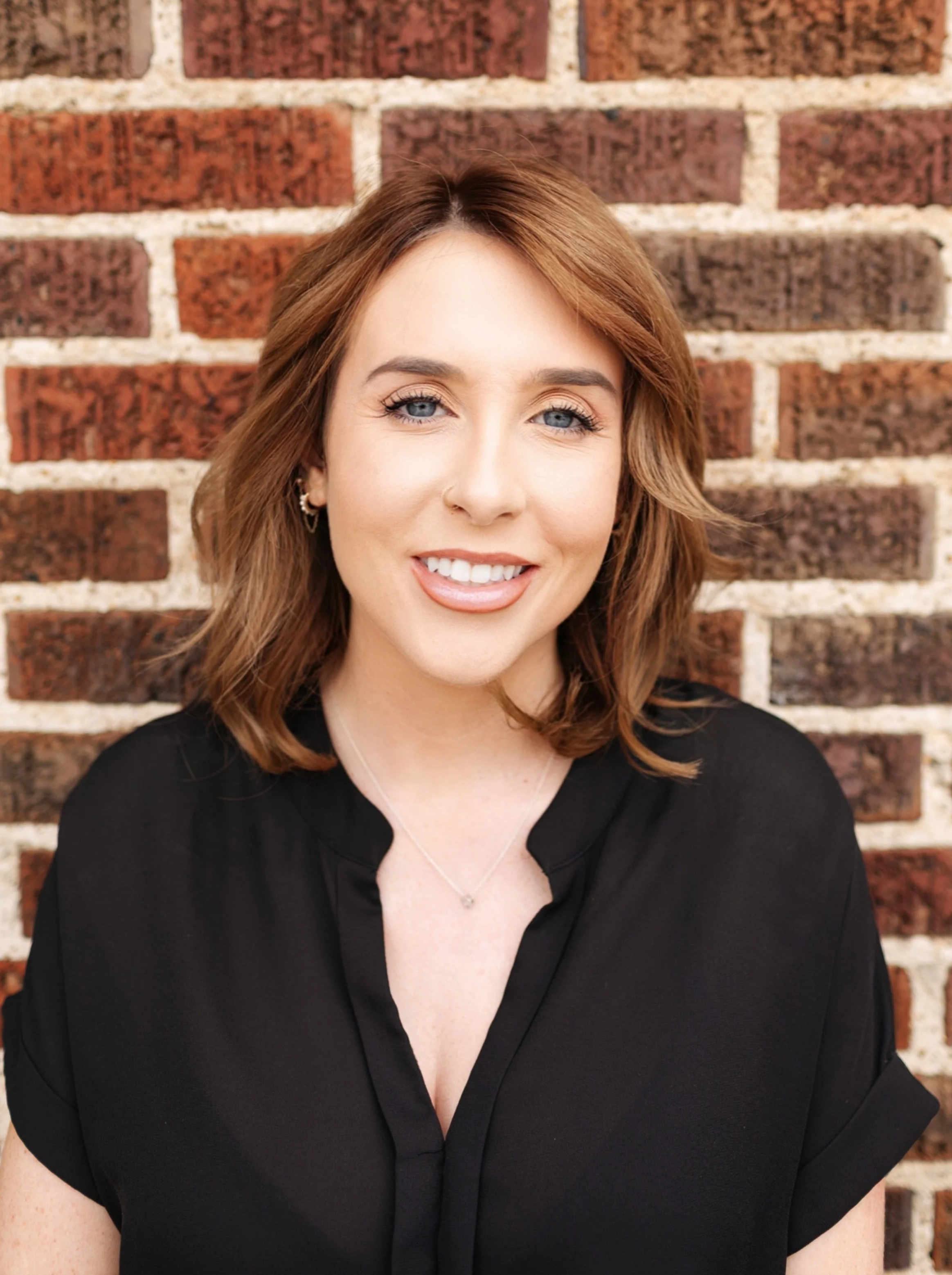 Close-up of a smiling woman with shoulder-length brown hair, wearing earrings, a necklace, and a black top, standing against a brick wall background.