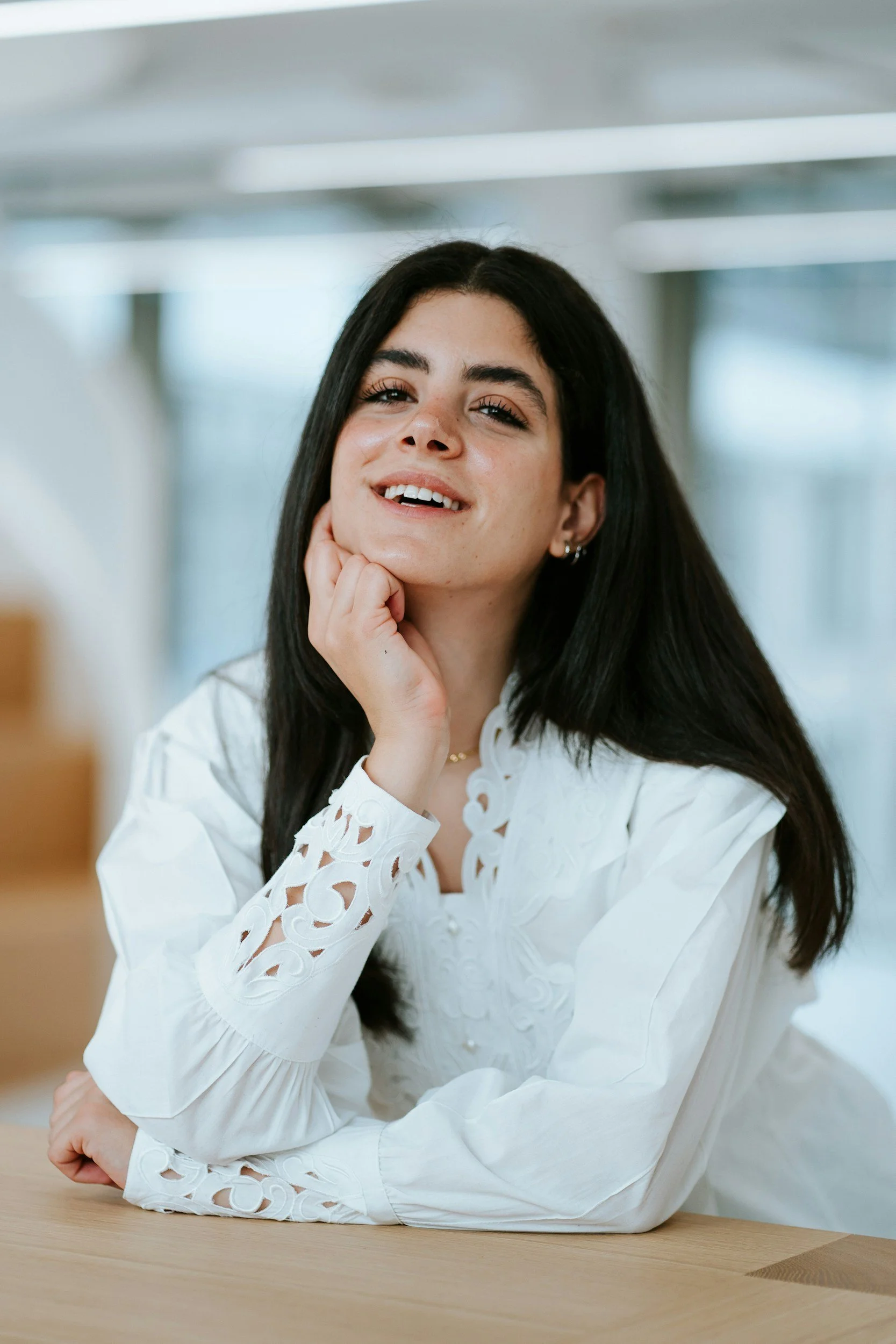 A woman with long dark hair wearing a white blouse with embroidered details, resting her chin on her hand and smiling at the camera, sitting at a table in a bright, modern interior.