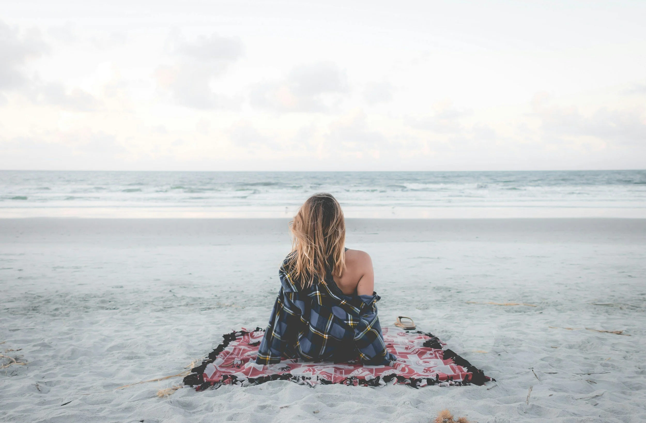 A person with long blonde hair sitting on a pink and black blanket on the sandy beach, facing the ocean with cloudy sky.