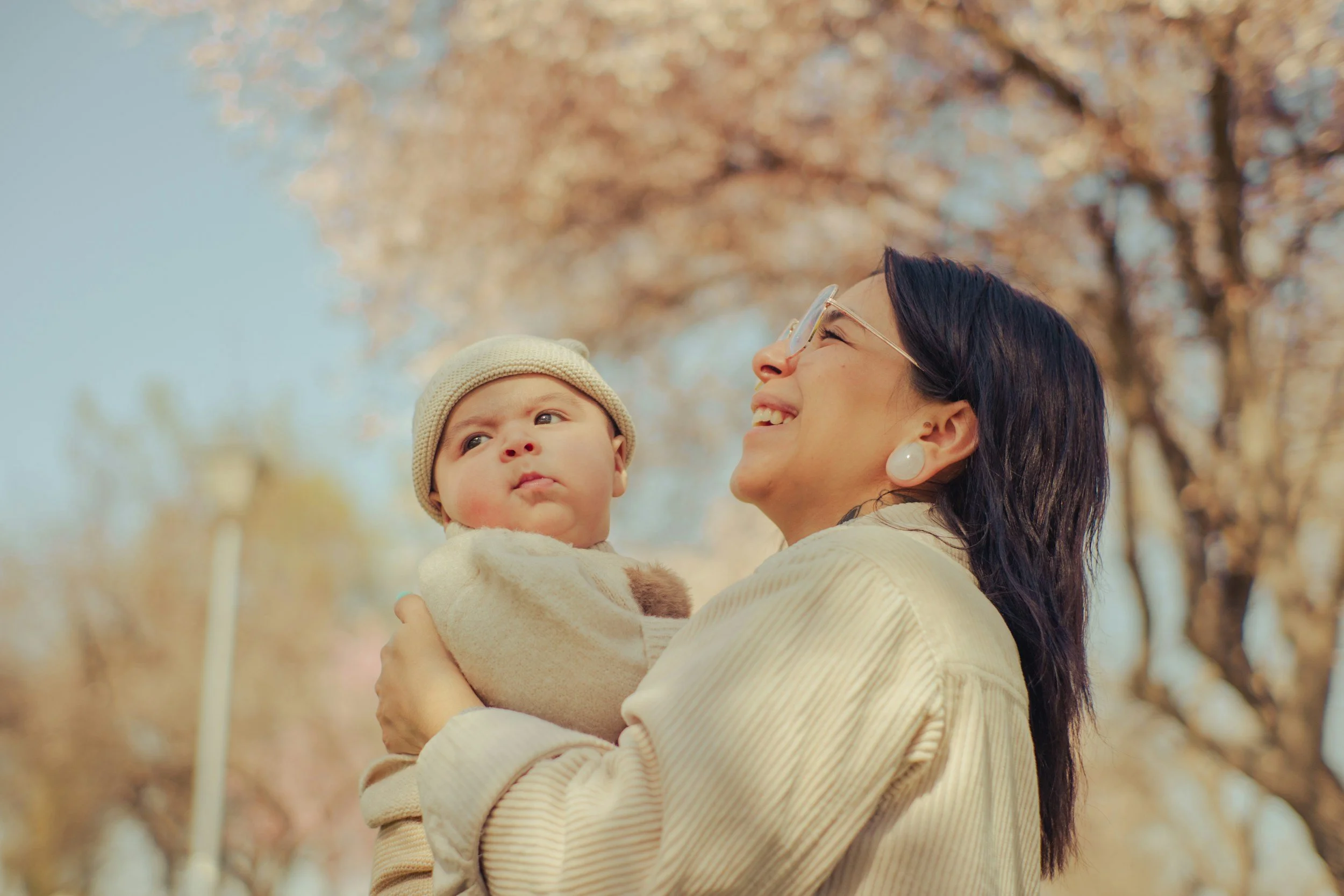 A woman with dark hair, glasses, and earrings holds a baby wearing a beige knit hat and cream-colored sweater outdoors, with trees and fall foliage in the background.