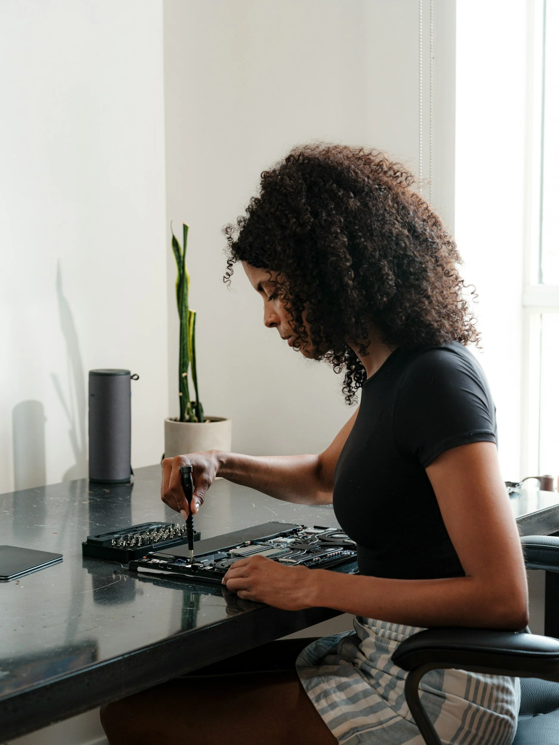 Woman with curly hair working with electronic components at a desk with a plant and smart speaker in a bright room.