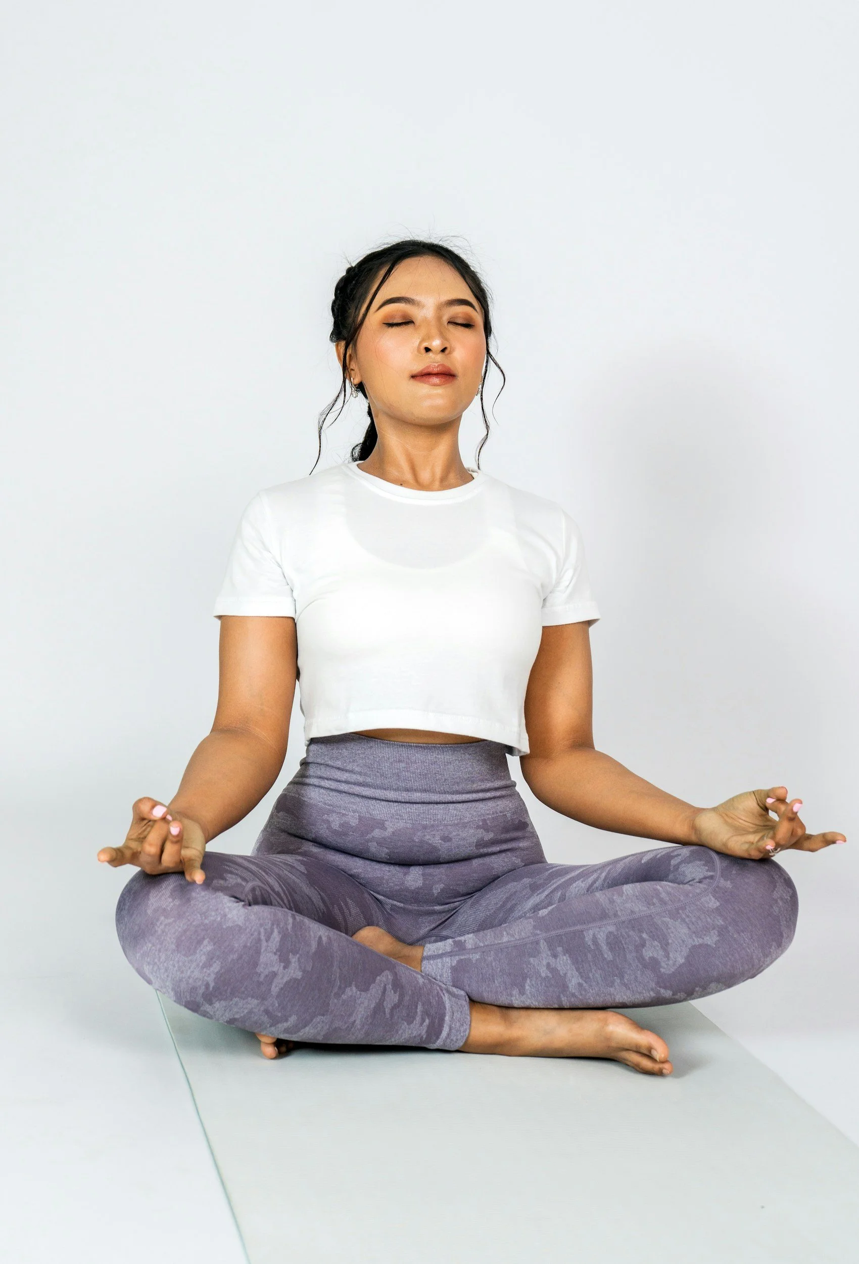 Woman practicing yoga in a seated cross-legged position, meditating with eyes closed, wearing a white t-shirt and purple camo-patterned leggings in a bright room.