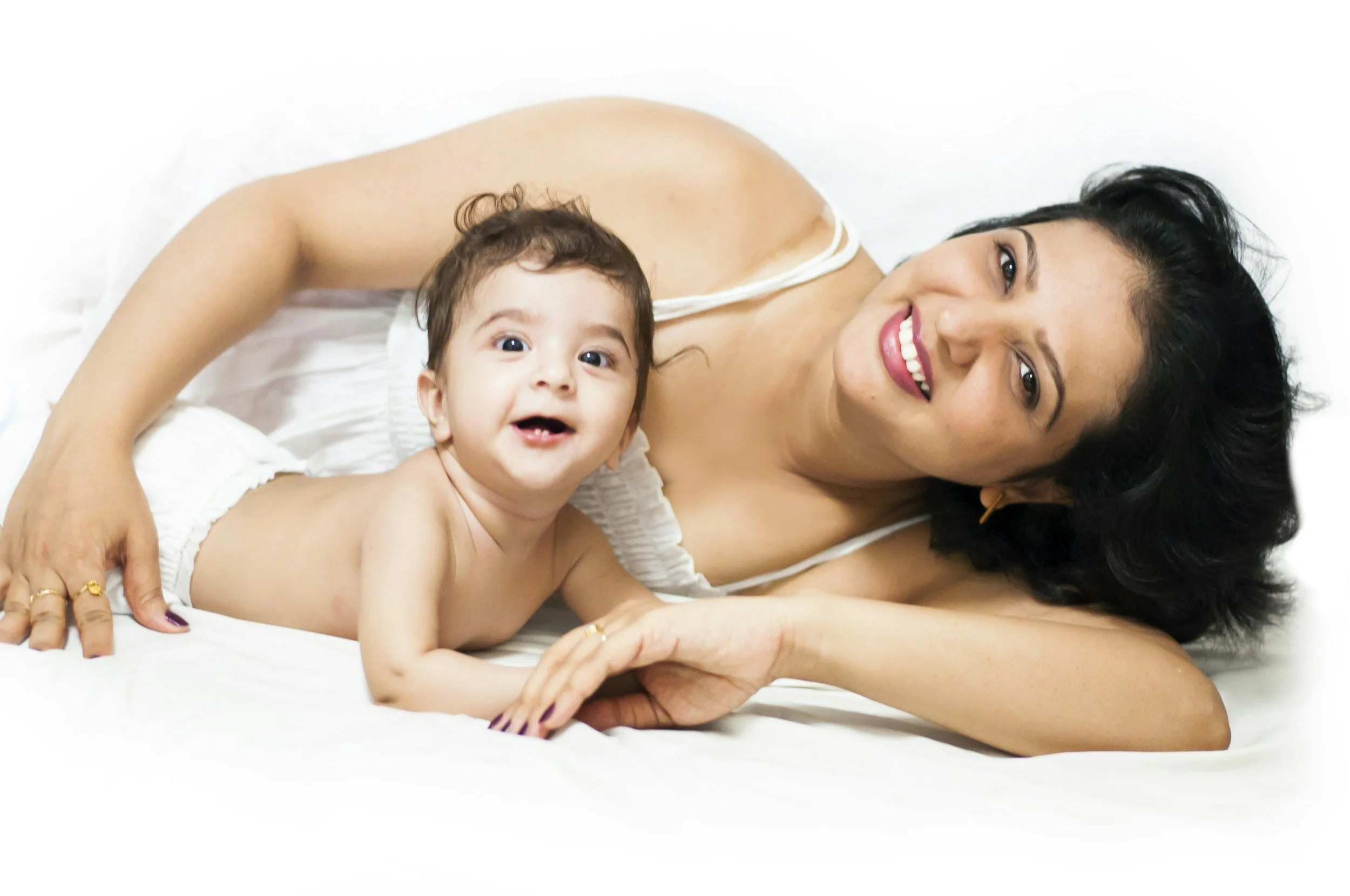 A woman with black hair and a bright smile lying on a white bed with a young child, both looking at the camera.