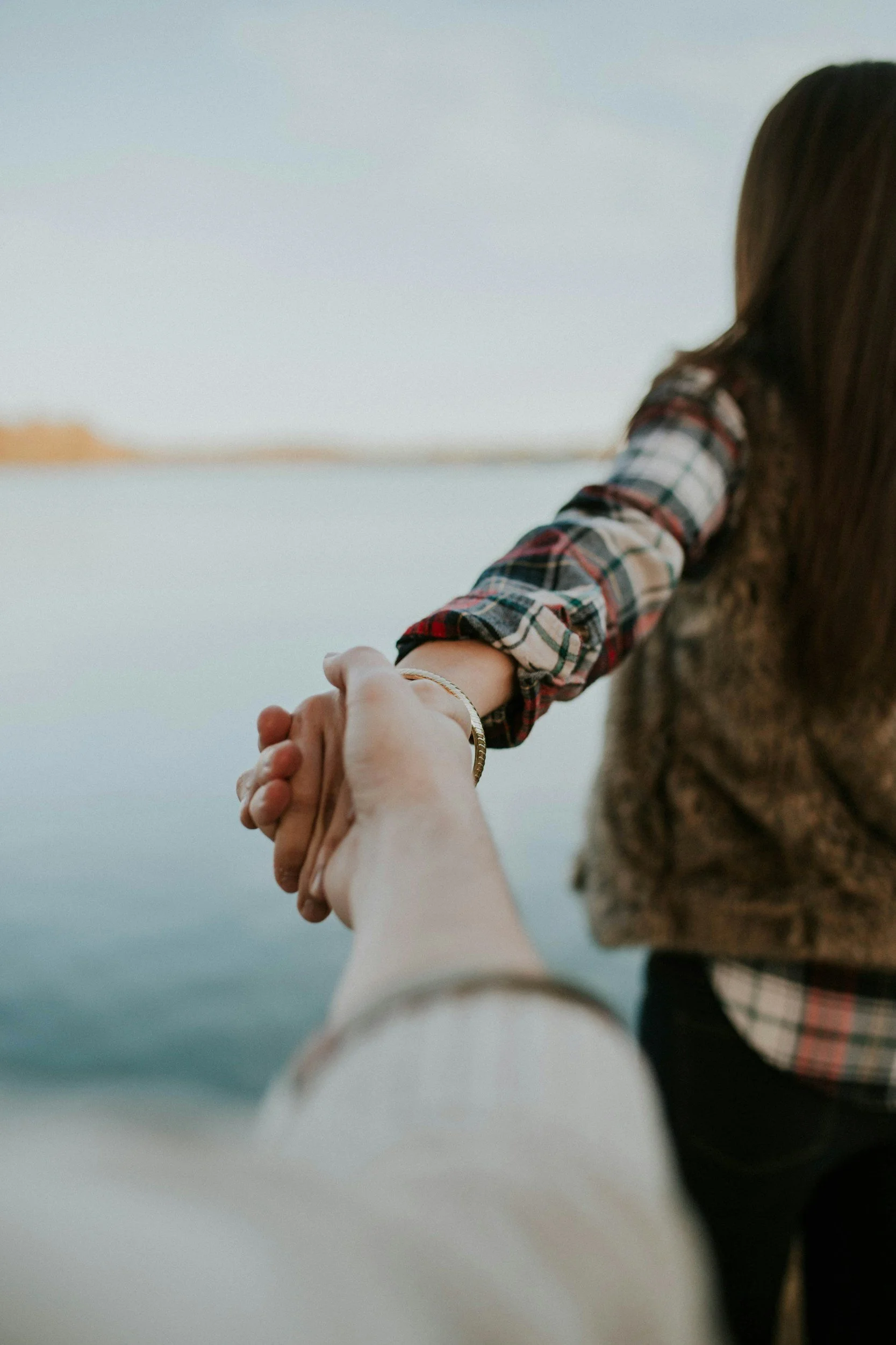 A person holding hands with a child near a body of water, with the child's arm and hand reaching out and the person's hand holding the child's hand.