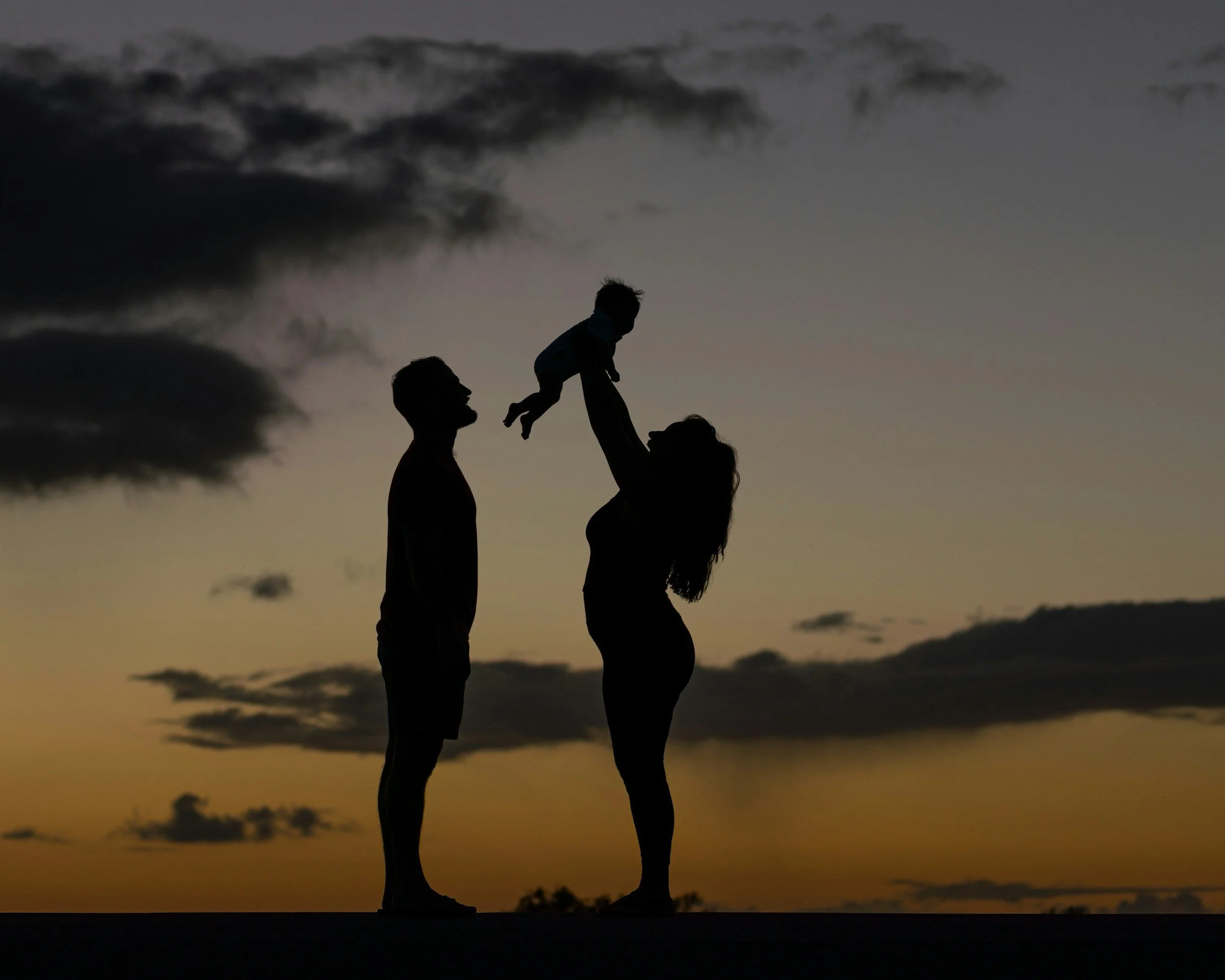 Silhouettes of a man, woman, and child outdoors during sunset, with the child being lifted in the air by the woman, and clouds in the evening sky.